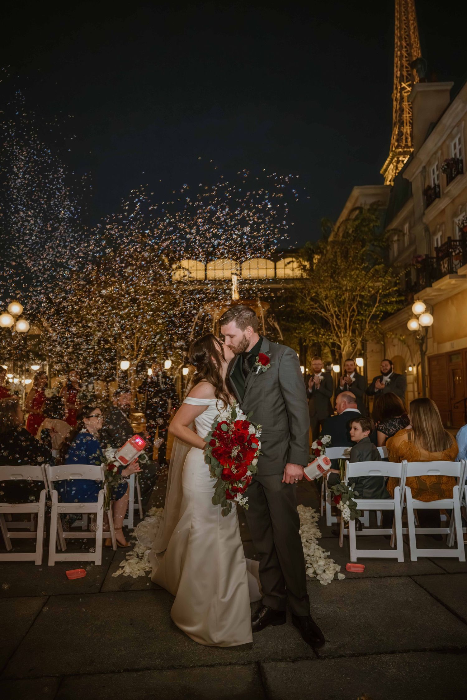 Bride and groom kissing at altar with confetti shower and Eiffel Tower backdrop at night.