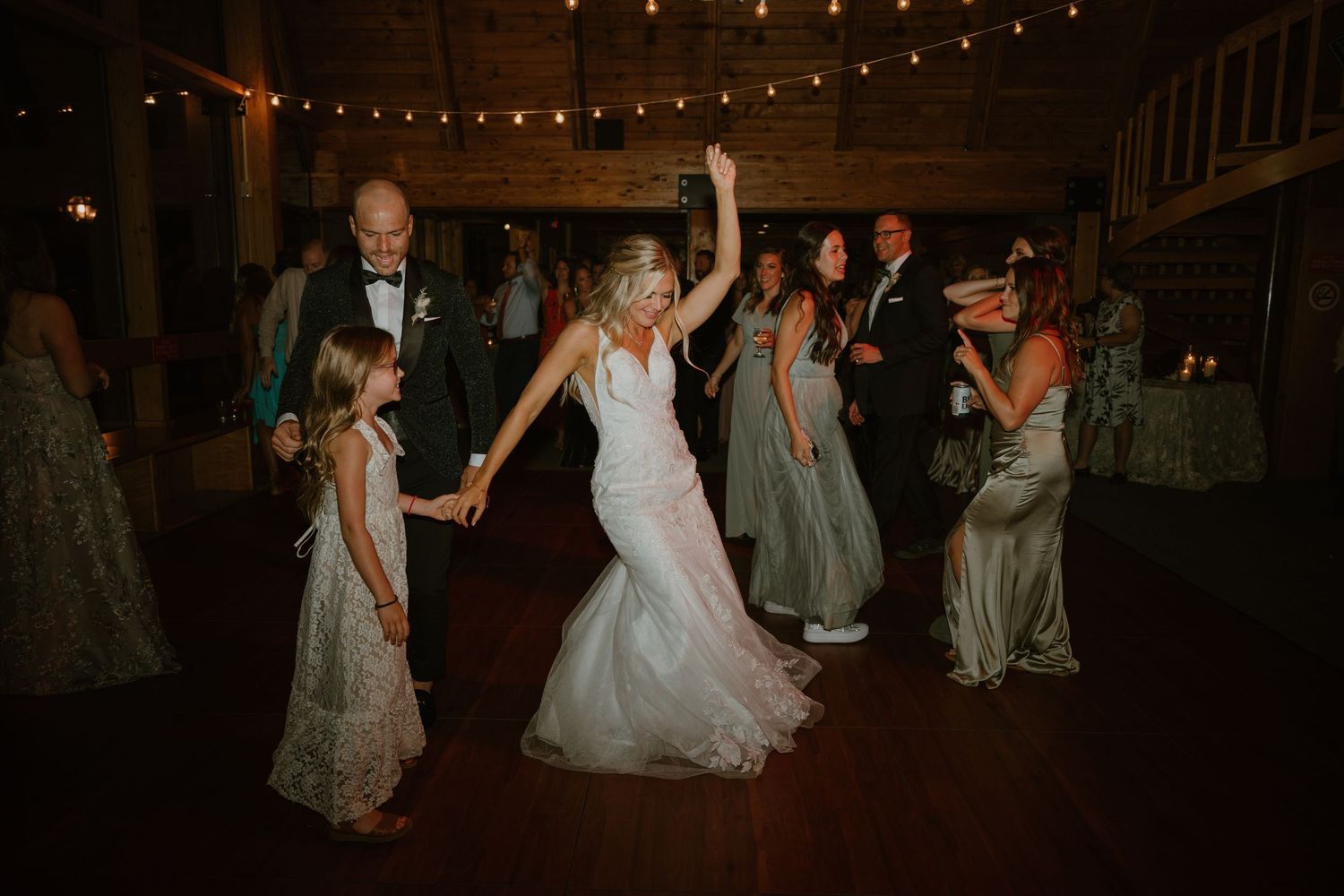 Bride in white gown dances joyfully with guests under string lights at reception.
