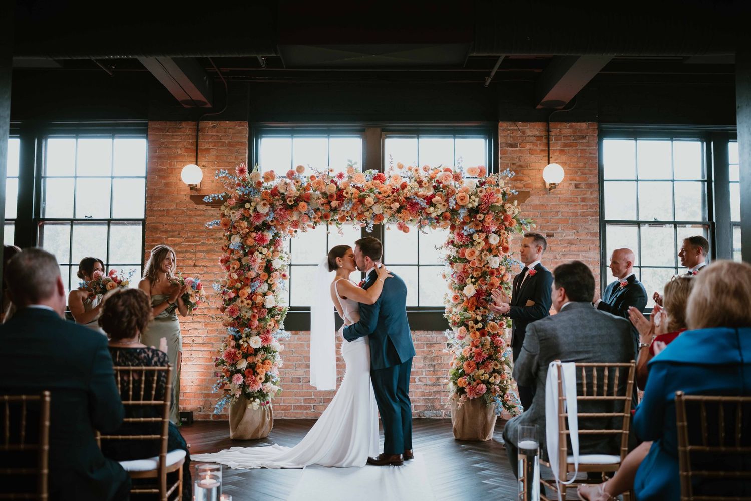 Bride and groom share first kiss under vibrant floral arch at altar.