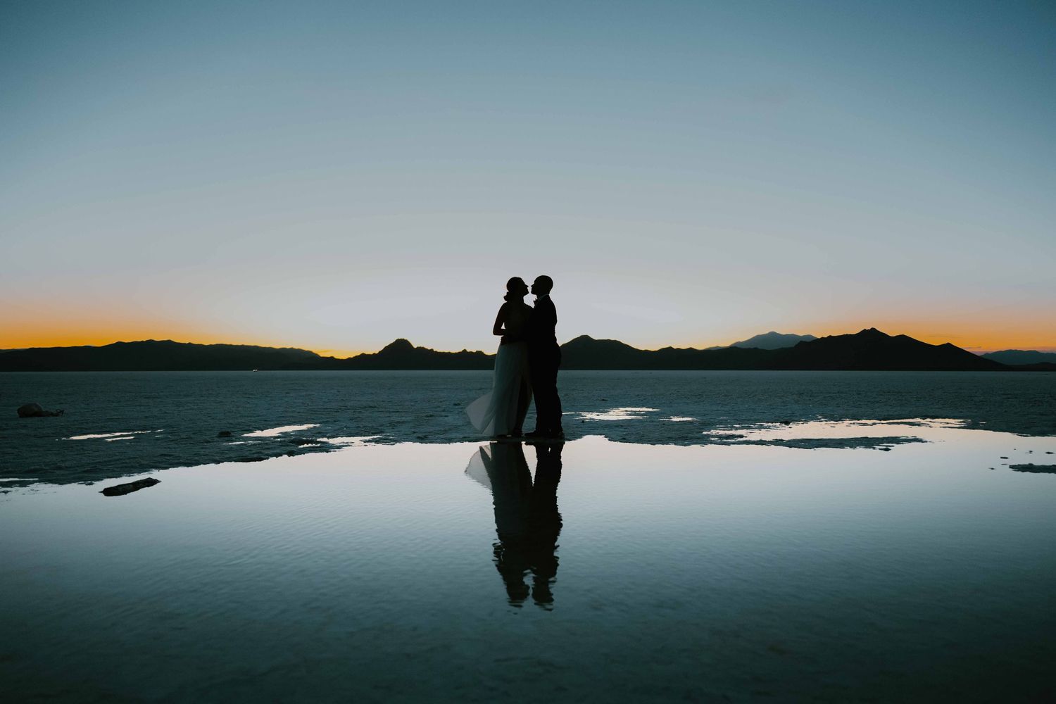 Silhouette of bride and groom reflected on salt flats at sunset with mountains.