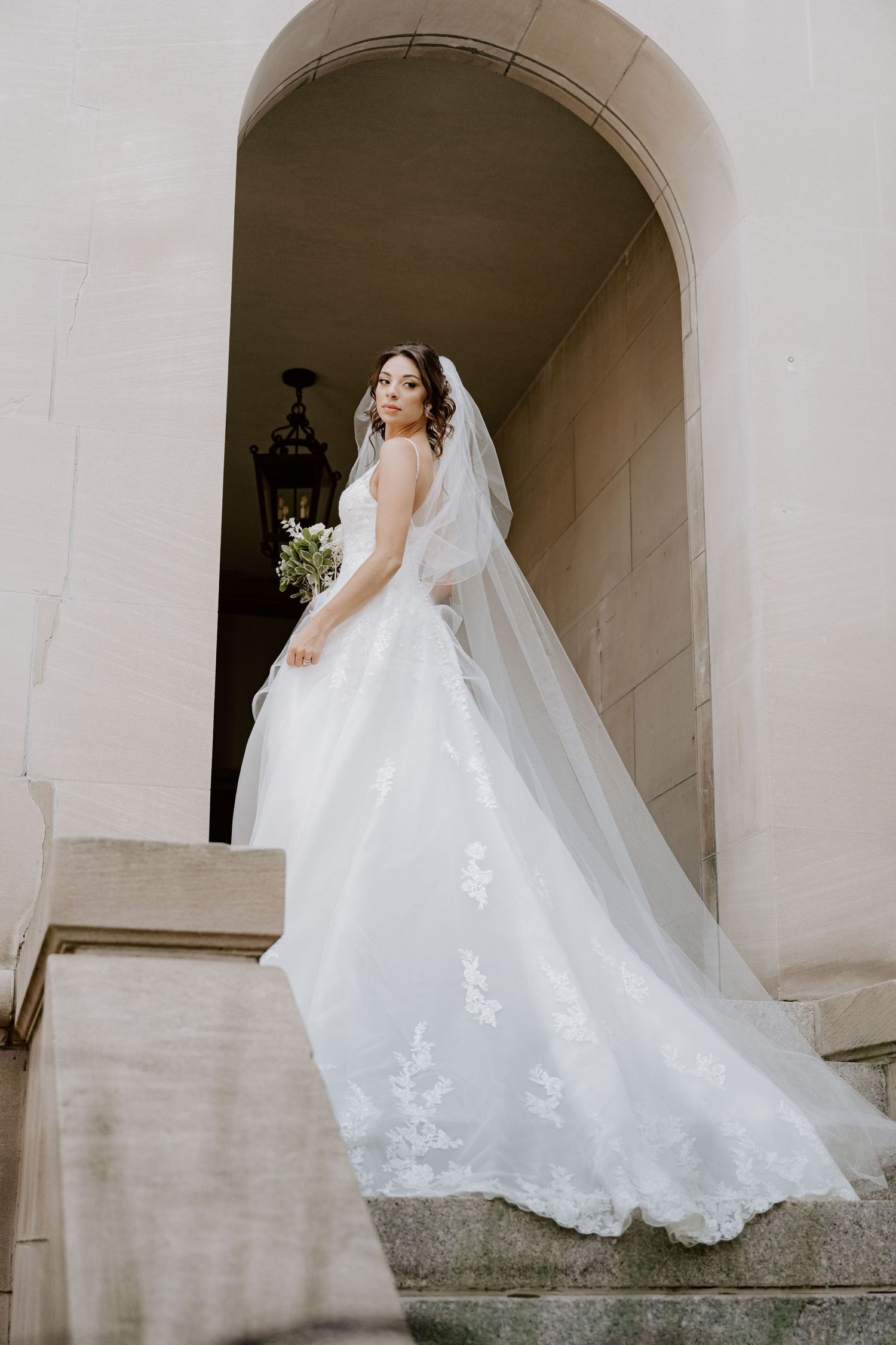 Elegant bride in lace ball gown with long veil holding bouquet beneath stone archway on steps.