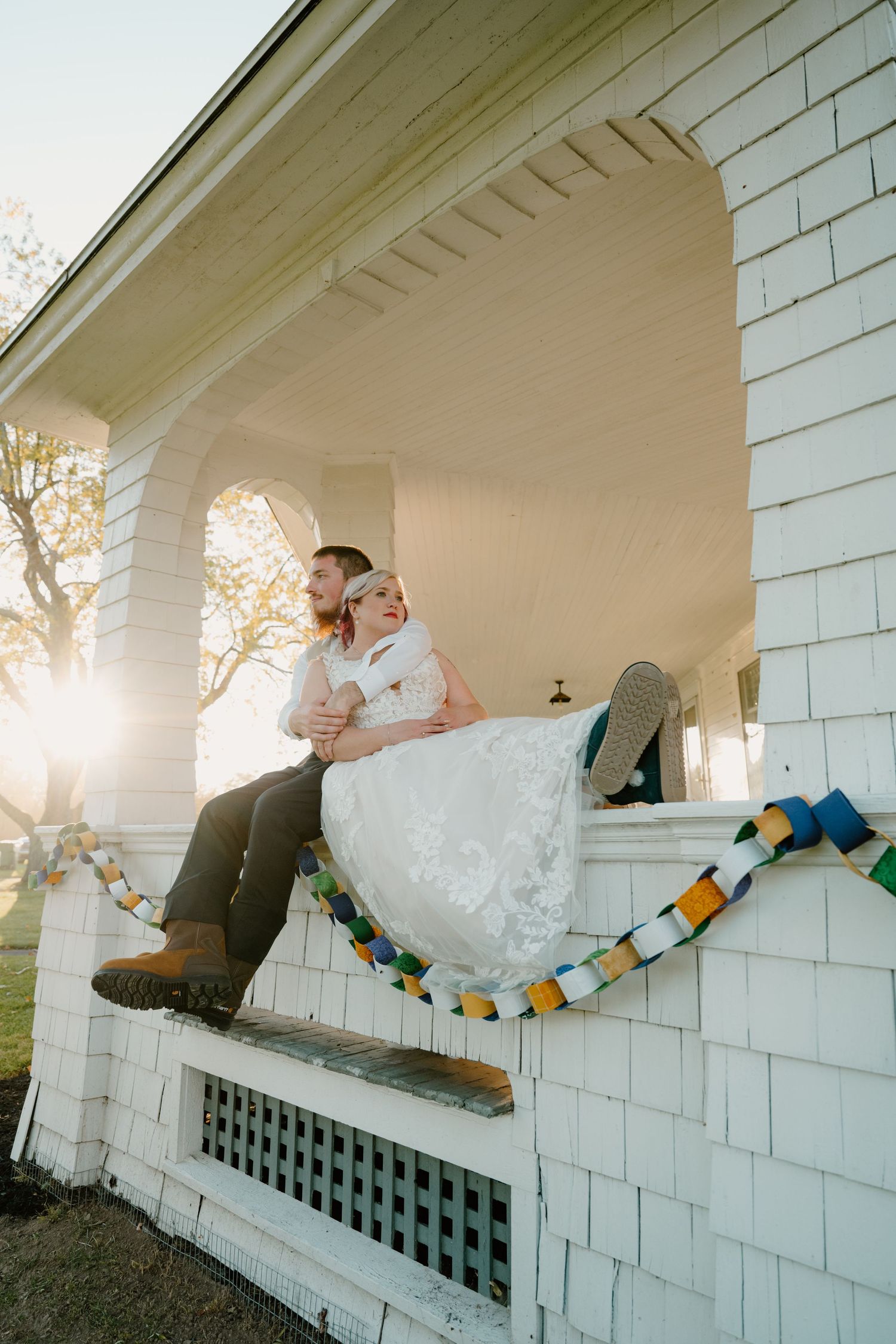 Bride and groom sit embracing on white cottage porch railing at golden sunset with colorful garland.