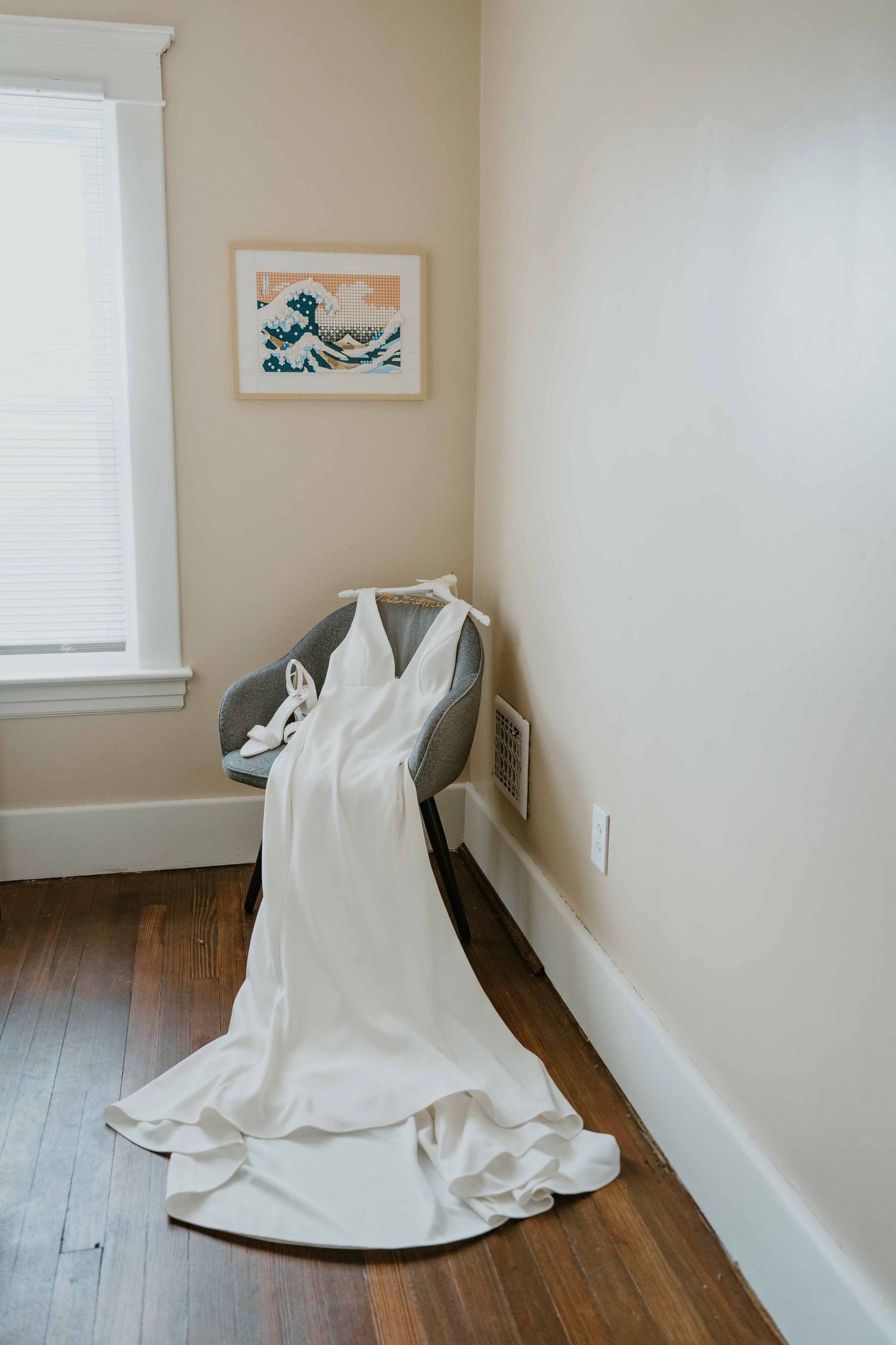 White wedding gown draped over a chair in a softly lit room with Hokusai wave art print.