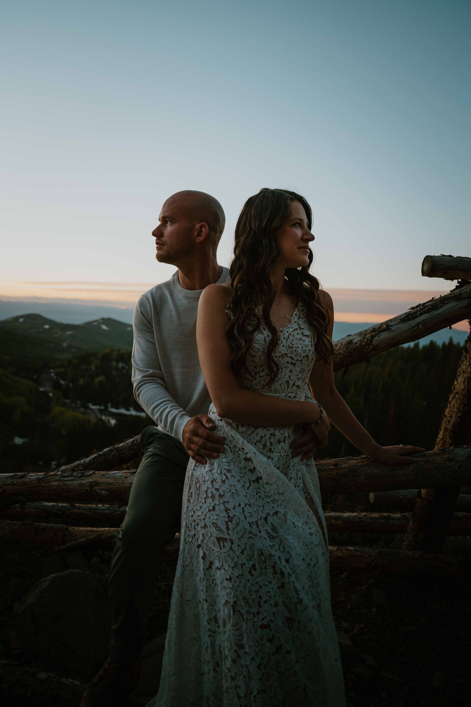 Romantic couple leaning against rustic log fence at dusk with scenic mountain backdrop.
