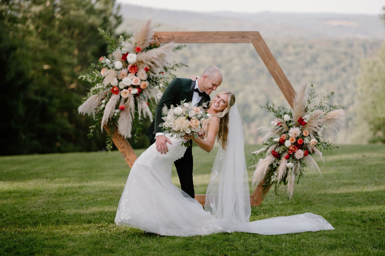 Bride and groom kissing under a hexagonal floral arch with pink and red blooms on a lush green hillside.