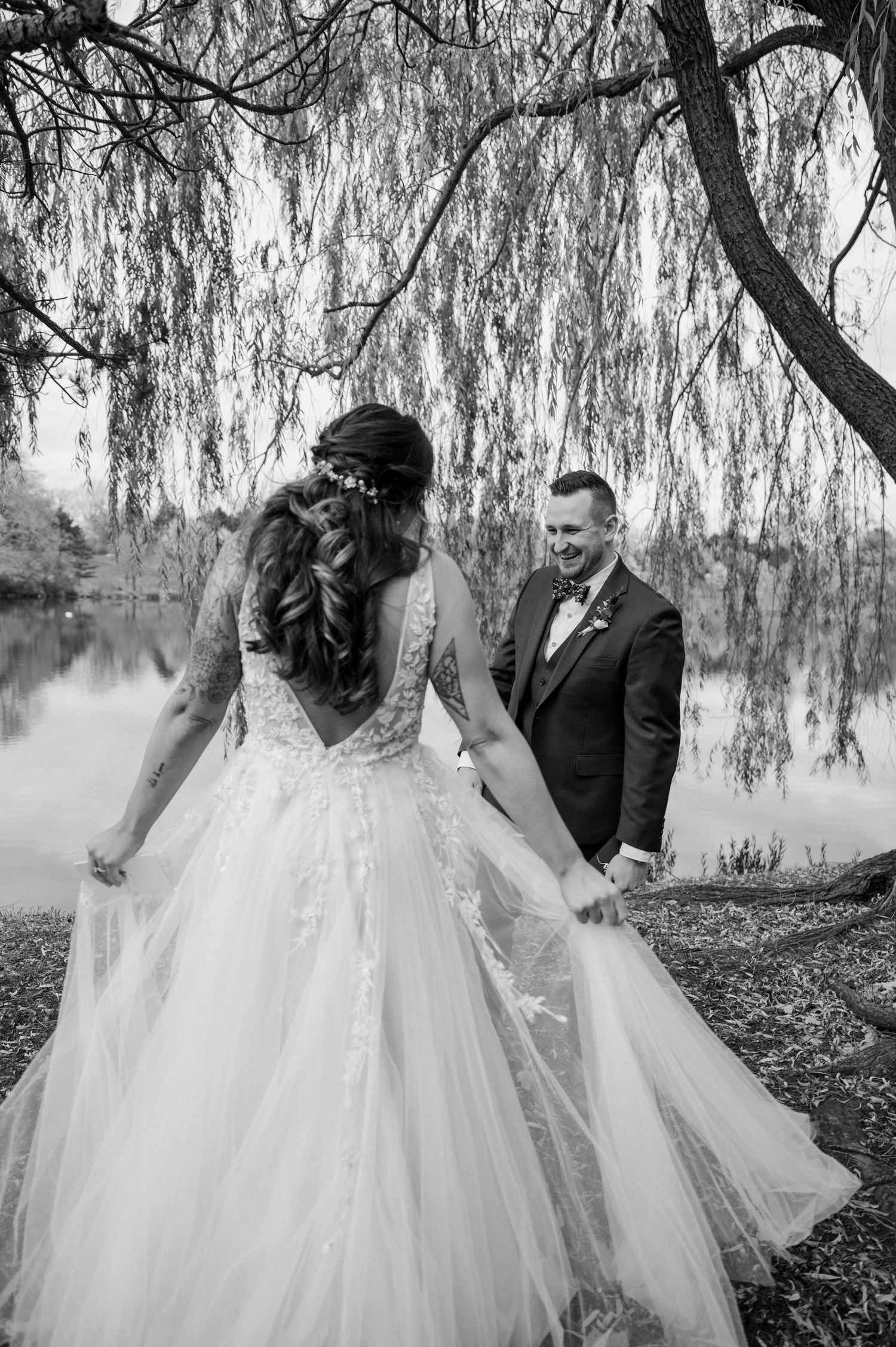 Black and white photo of bride and groom holding hands under weeping willow tree by lake.