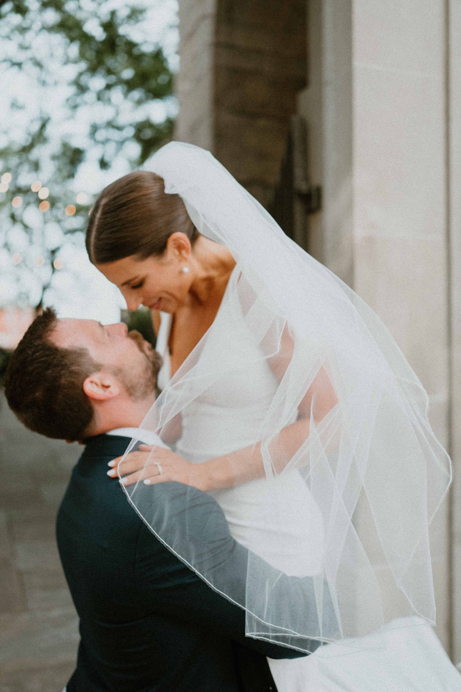 Groom lifts smiling bride in a white gown and veil for a joyful moment at an outdoor wedding venue.