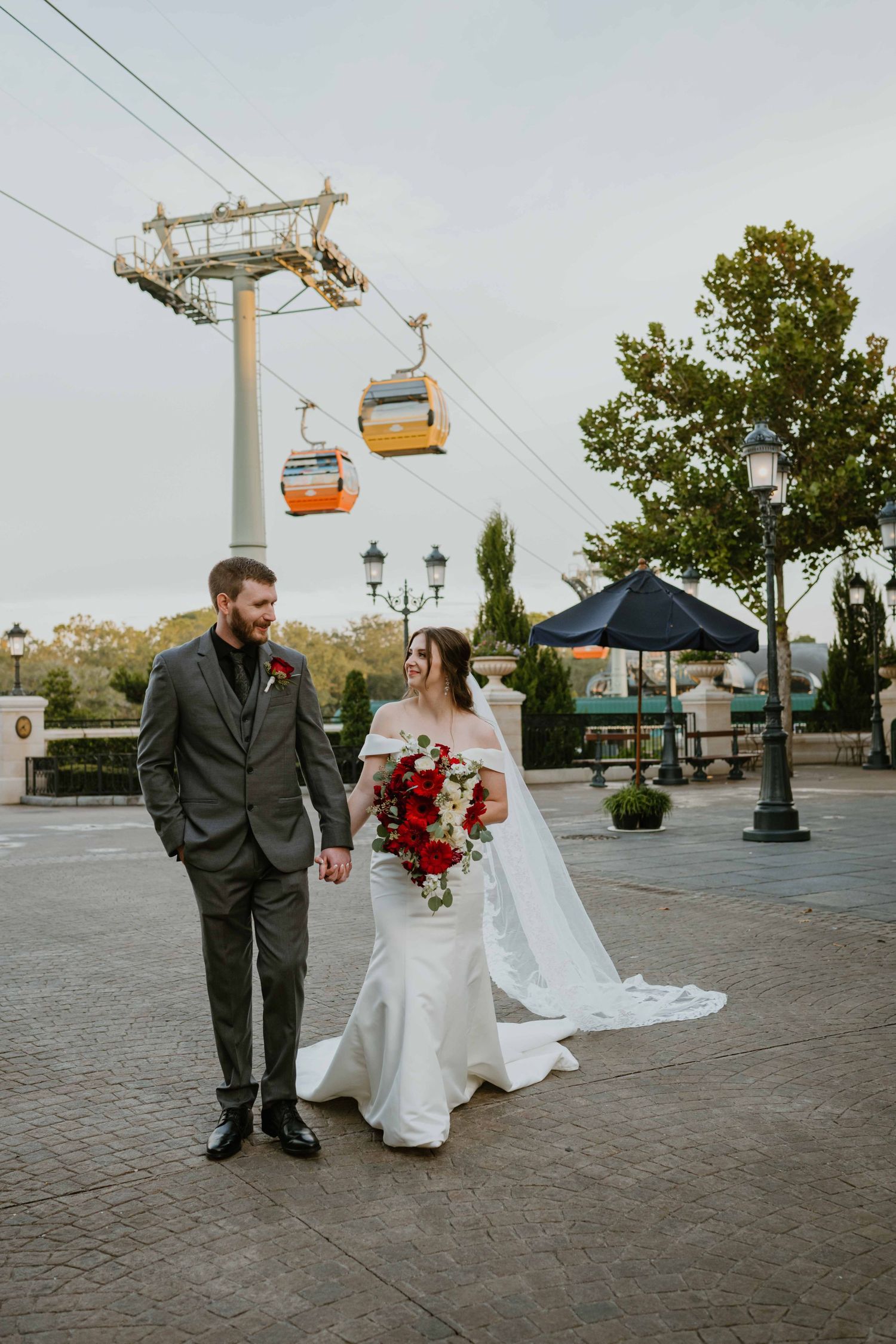 Bride and groom walk hand in hand beneath Disney Skyliner gondolas during their Epcot wedding portraits.
