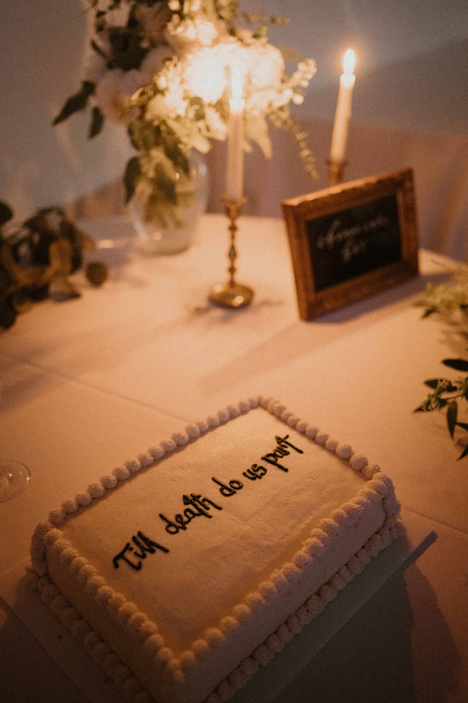 Wedding sheet cake reads Till death do us part by candlelight on a reception table.