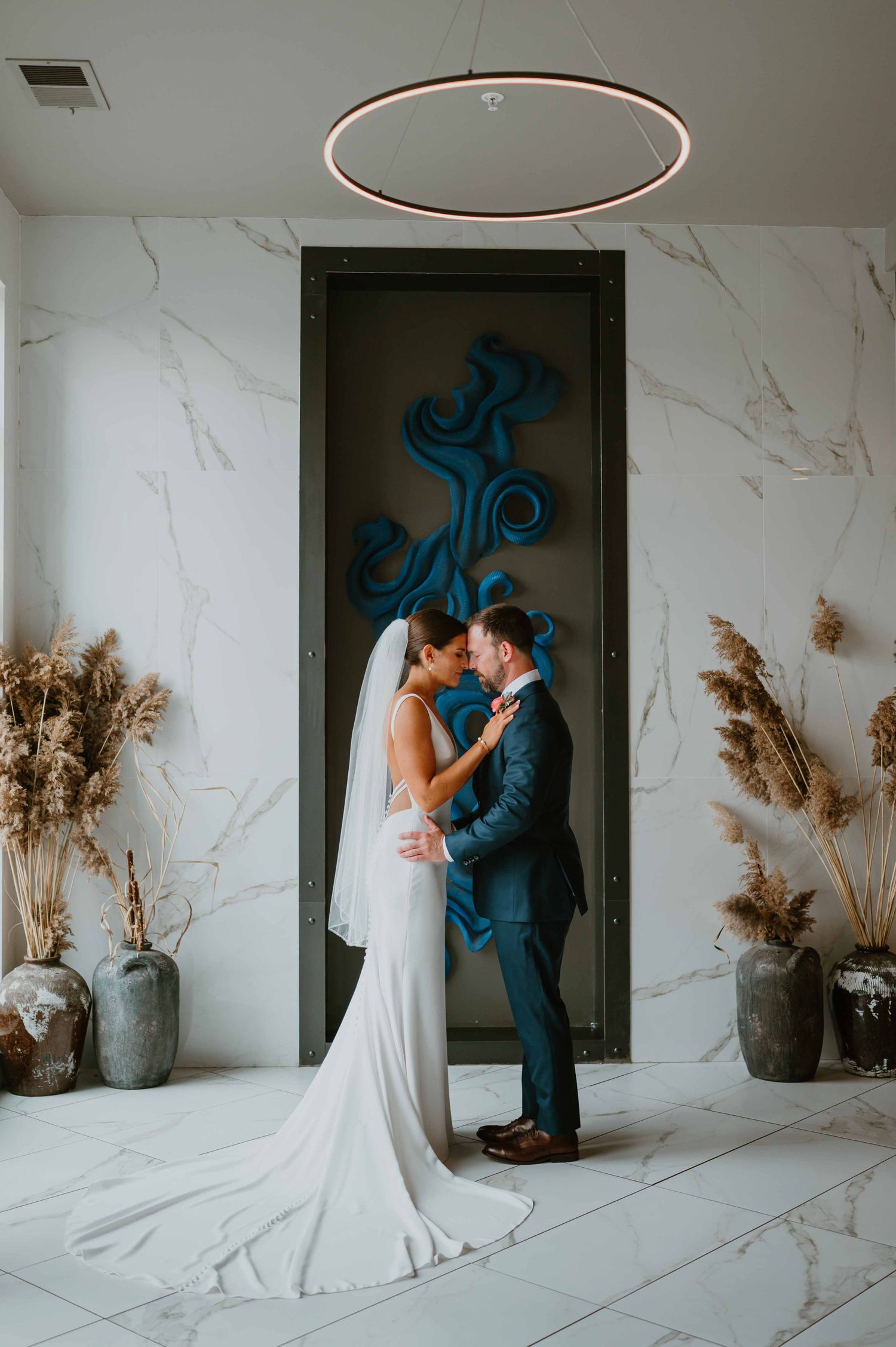 Bride and groom share a romantic moment in a modern marble venue with blue decor and dried pampas grass arrangements.