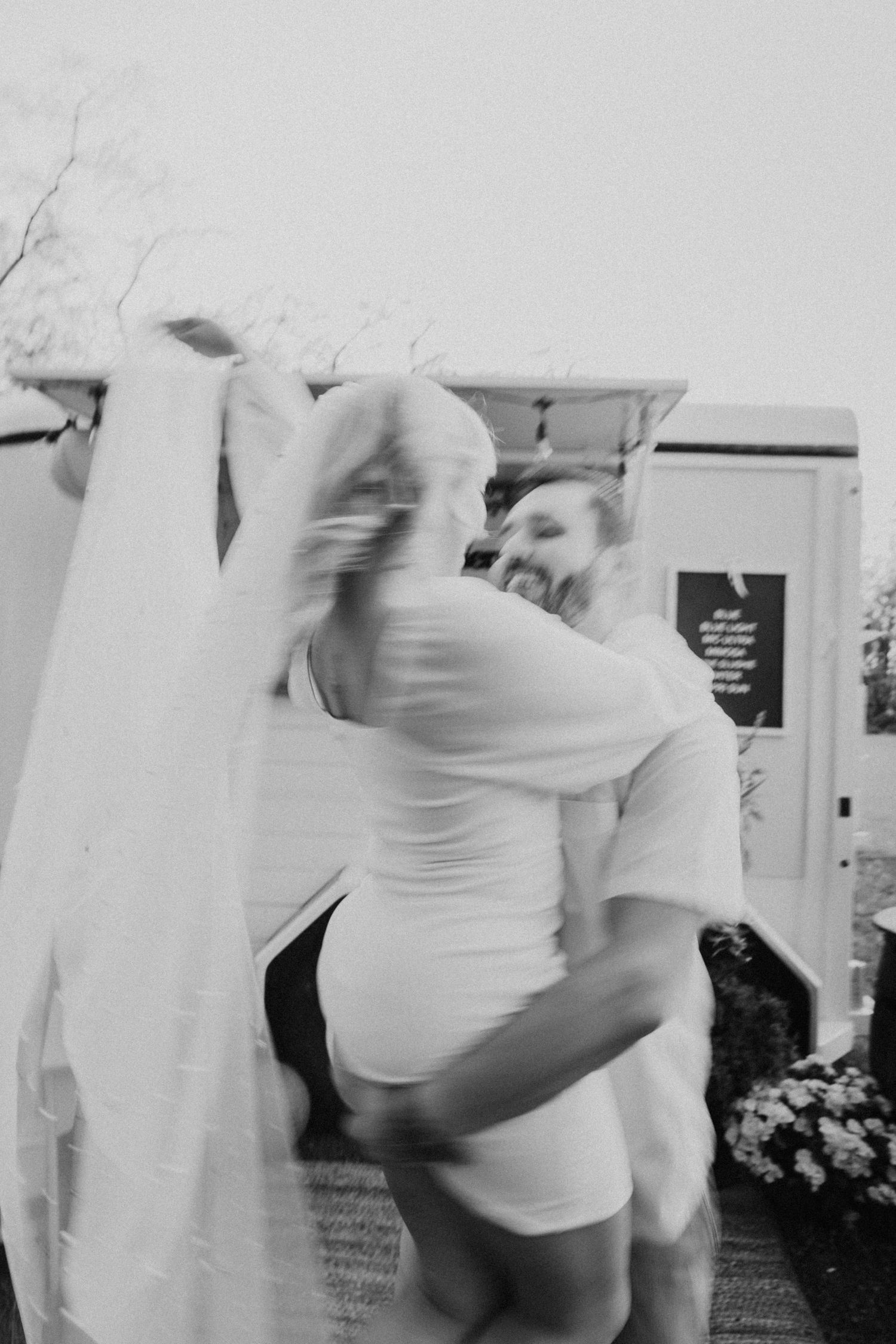 Black and white photo of groom lifting bride in wedding attire outside a white food truck.