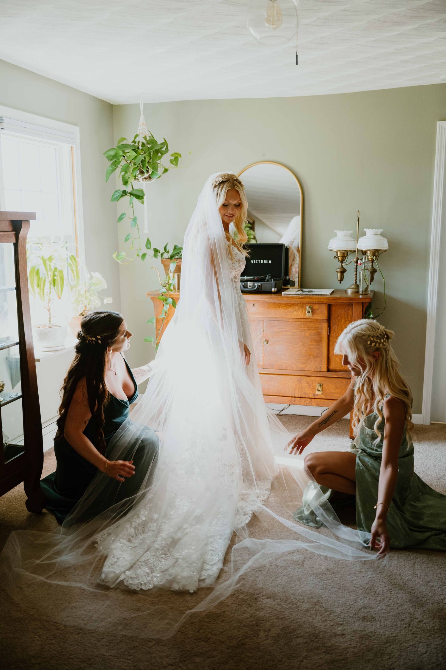 Two bridesmaids in green dresses arranging bride's cathedral veil on floor before wedding ceremony.