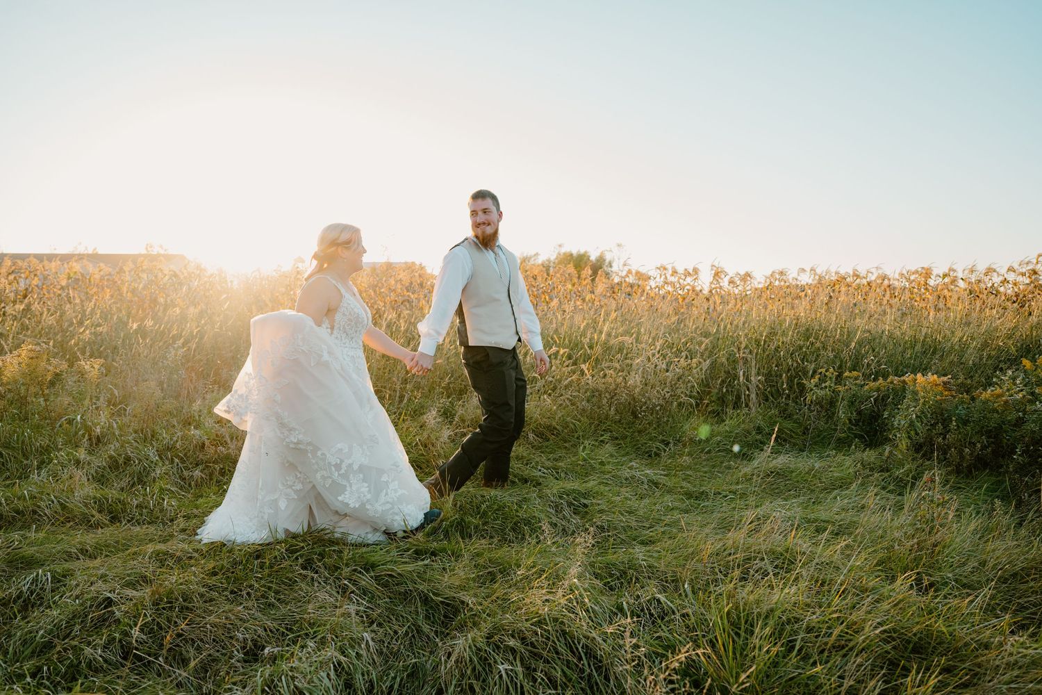 Bride and groom walk hand-in-hand through sunlit golden meadow grass during outdoor wedding celebration.