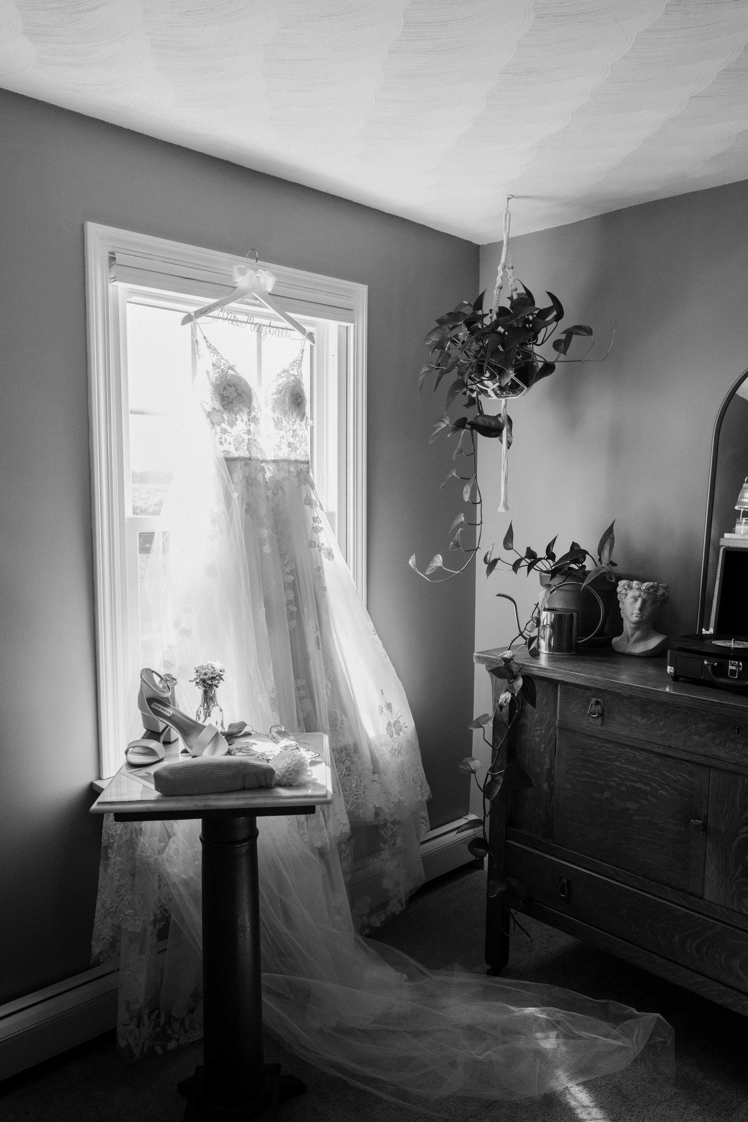 Black and white photo of lace wedding dress hanging in window with shoes and antique dresser.
