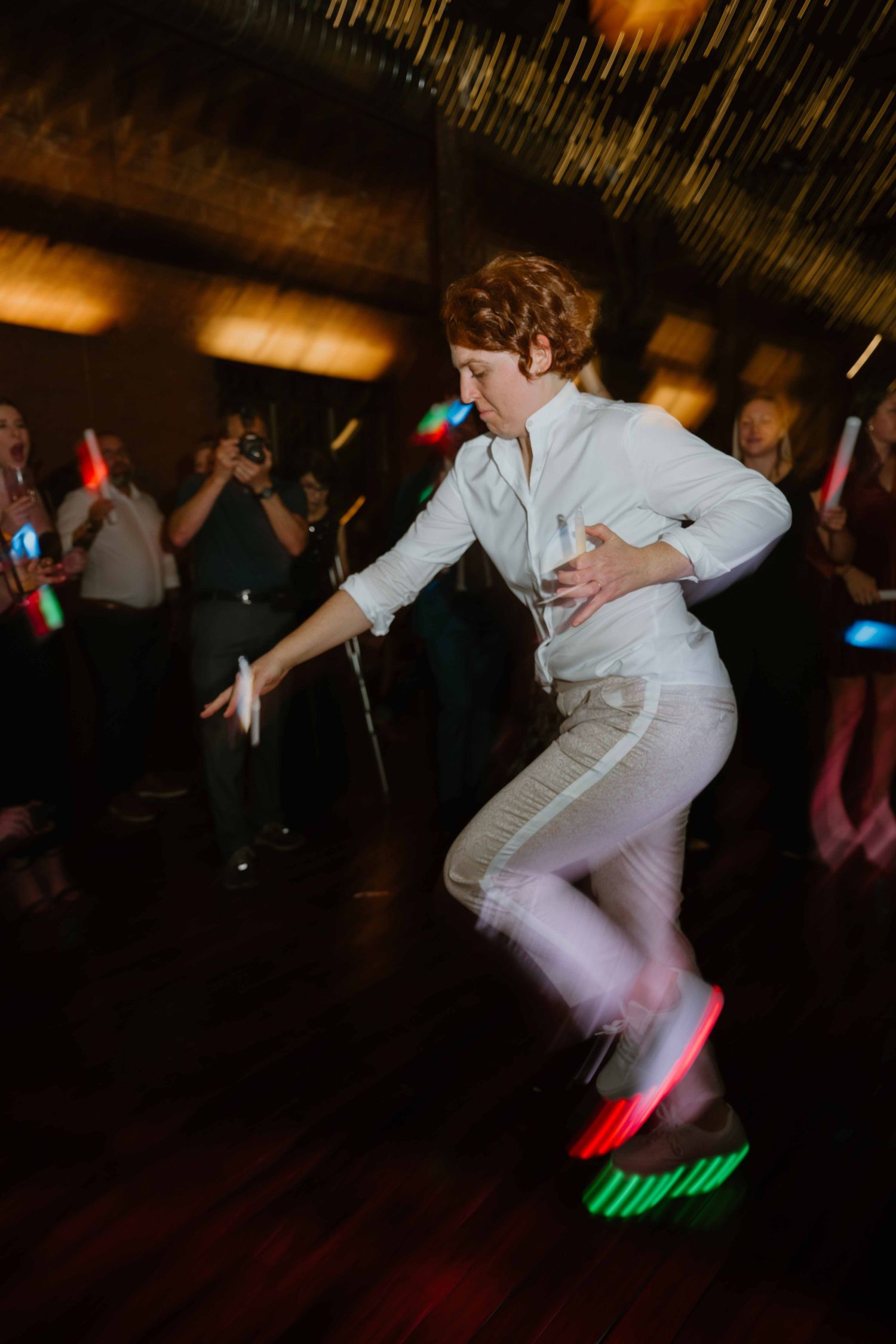 Energetic wedding guest in white suit dances with LED light sticks on a lively reception dance floor.