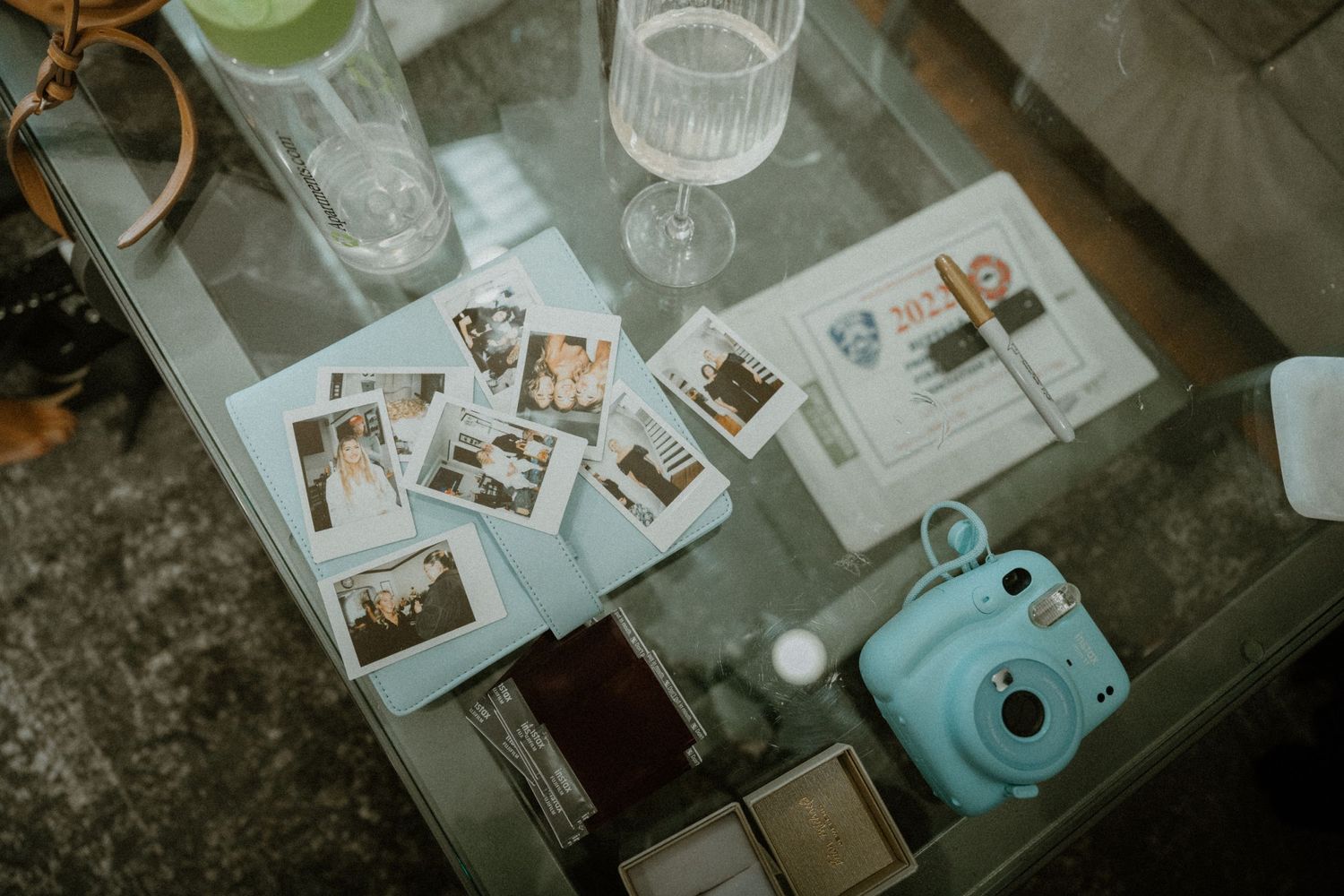 Flat lay of Polaroid photos, Instax Mini camera, cocktail glasses, and wedding stationery on glass table.