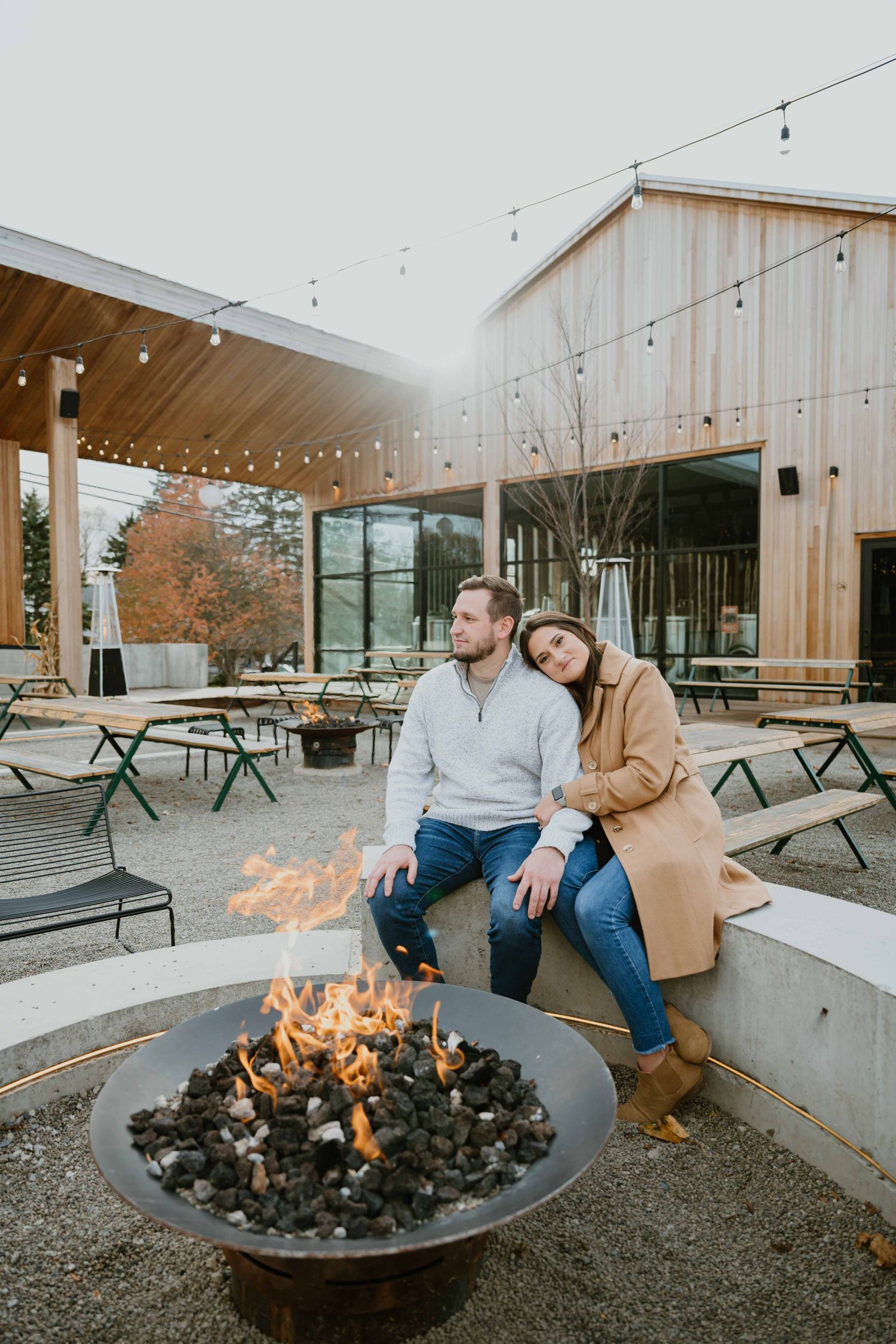 Couple relaxes by fire pit at modern outdoor venue with string lights overhead.