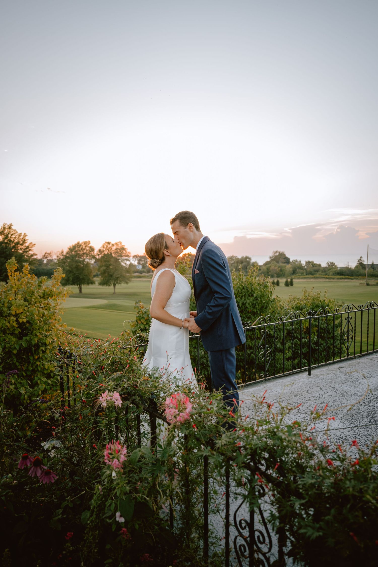 Bride and groom kissing at sunset on a garden terrace with pink flowers and a golf course backdrop.