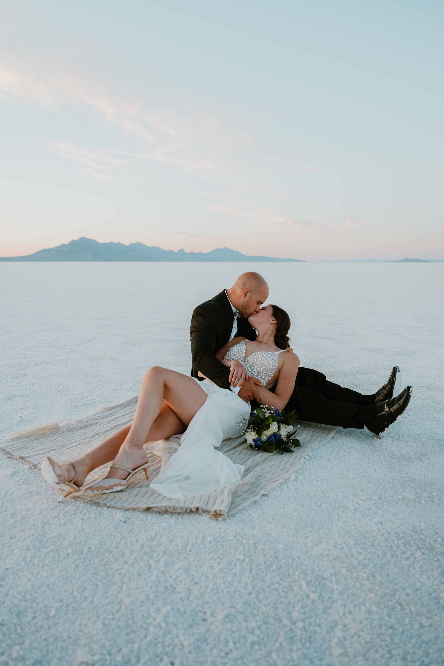 Groom leans in to kiss bride as she reclines on a blanket on the glowing white salt flats.