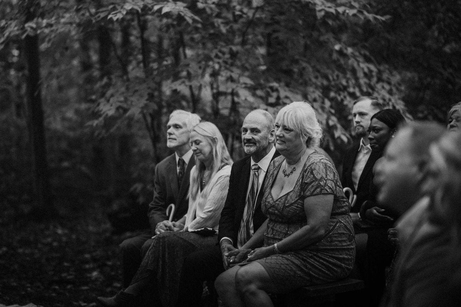 Guests seated on wooden benches watching an outdoor forest wedding ceremony in black and white.
