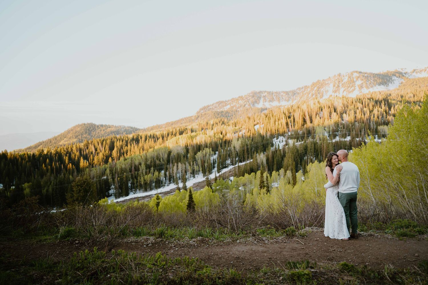 Wedding couple holds hands on mountain overlook with golden forest and snow-dusted peaks behind.