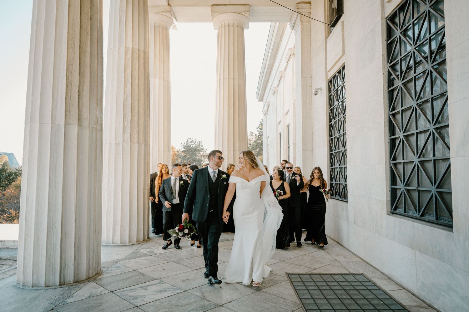 Bride and groom walk between grand columns with wedding party celebrating behind them at sunset.