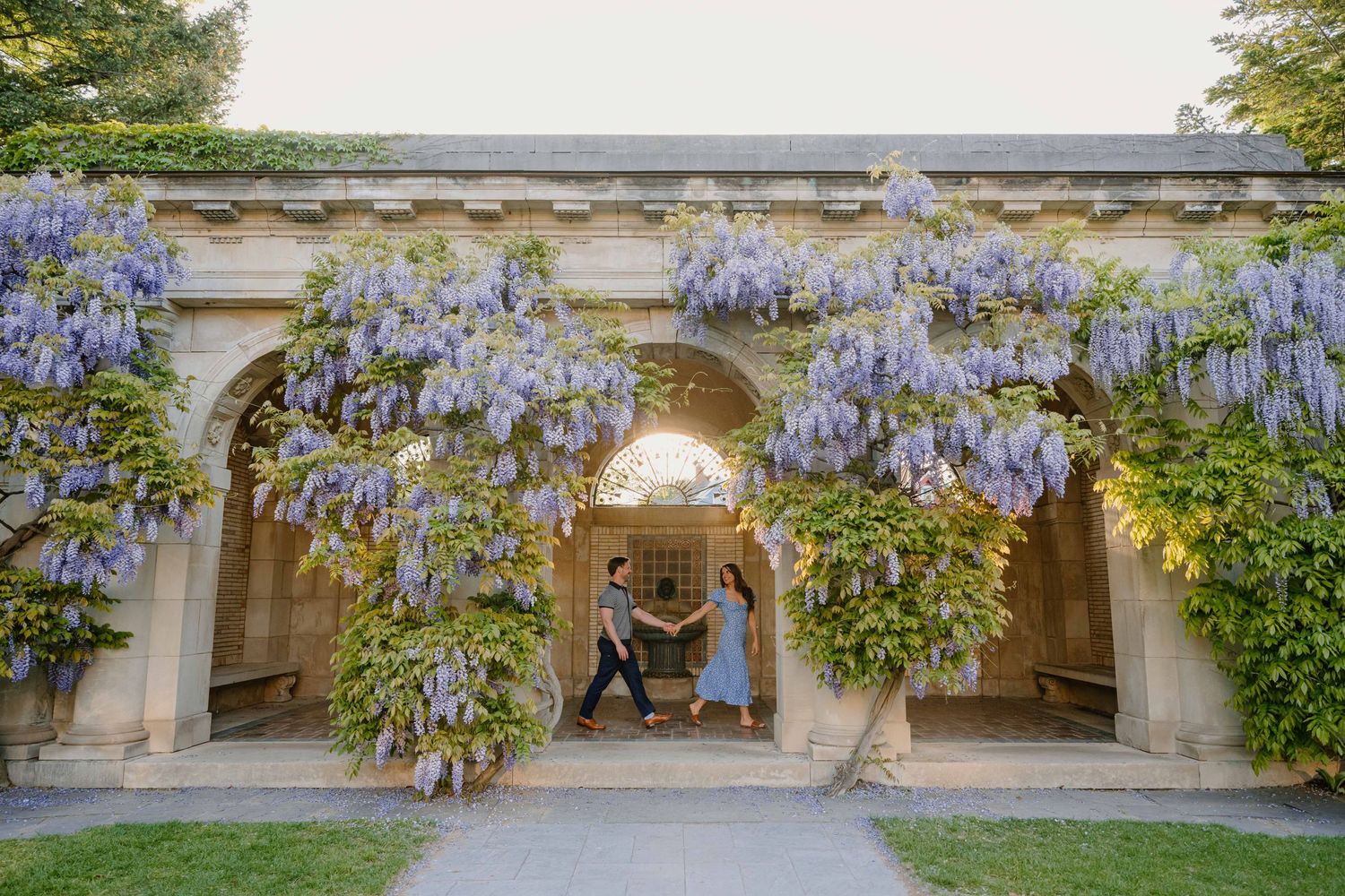 Couple dances playfully under stunning purple wisteria-covered stone archway at a historic garden estate.
