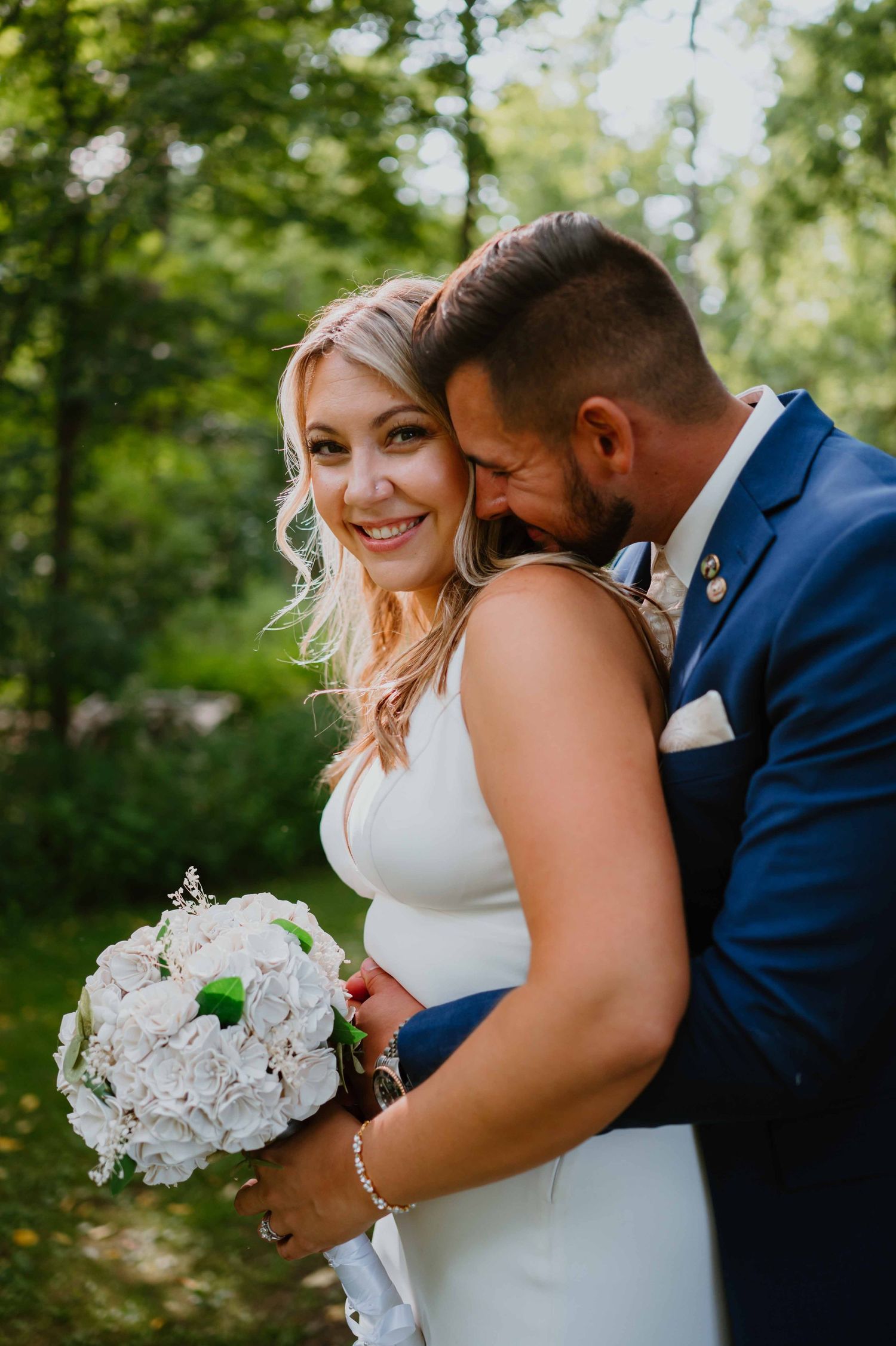 Smiling bride in white gown holding white bouquet while groom in navy suit embraces her outdoors.
