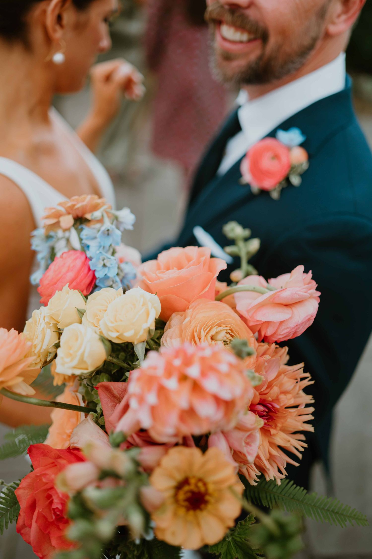 Close-up of vibrant wedding bouquet with peach, coral, and cream blooms held by smiling bride near groom in navy suit.