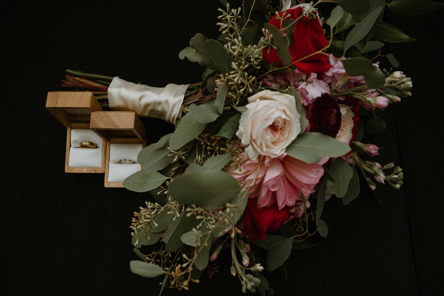 Wedding bouquet of red roses and pink blooms with gold velvet ring boxes displayed.