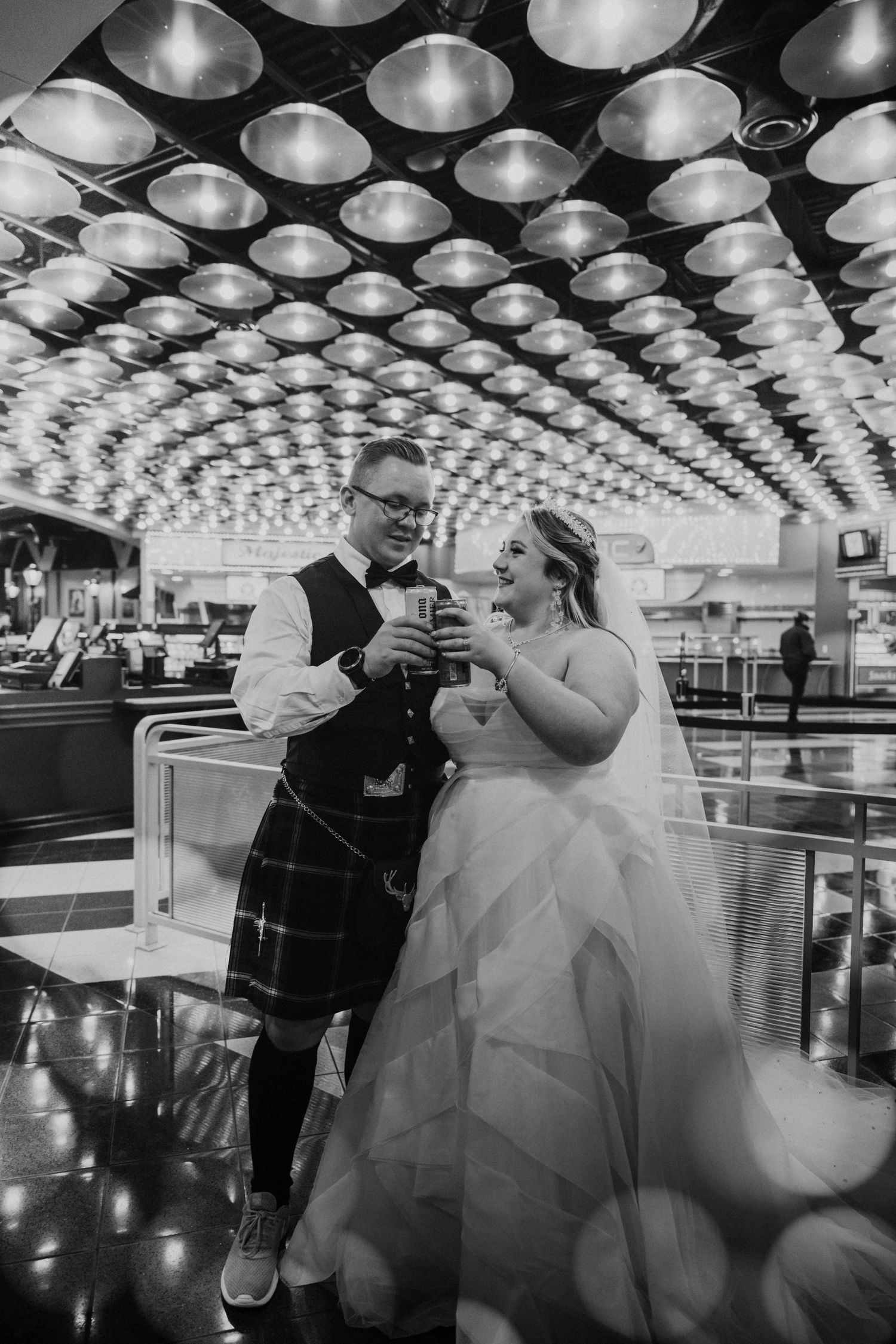 Black and white photo of bride and groom in kilt toasting drinks under geometric ceiling lights at Disney.