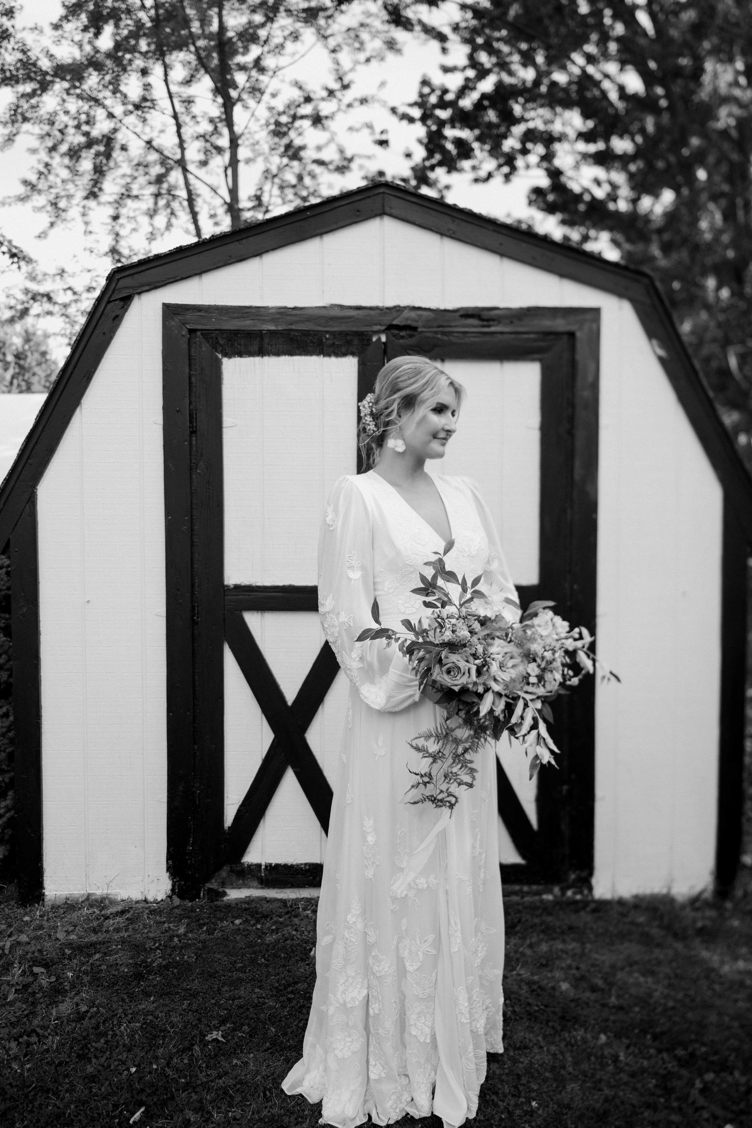 Bride in lace gown holding bouquet stands before a white barn in black and white photo.