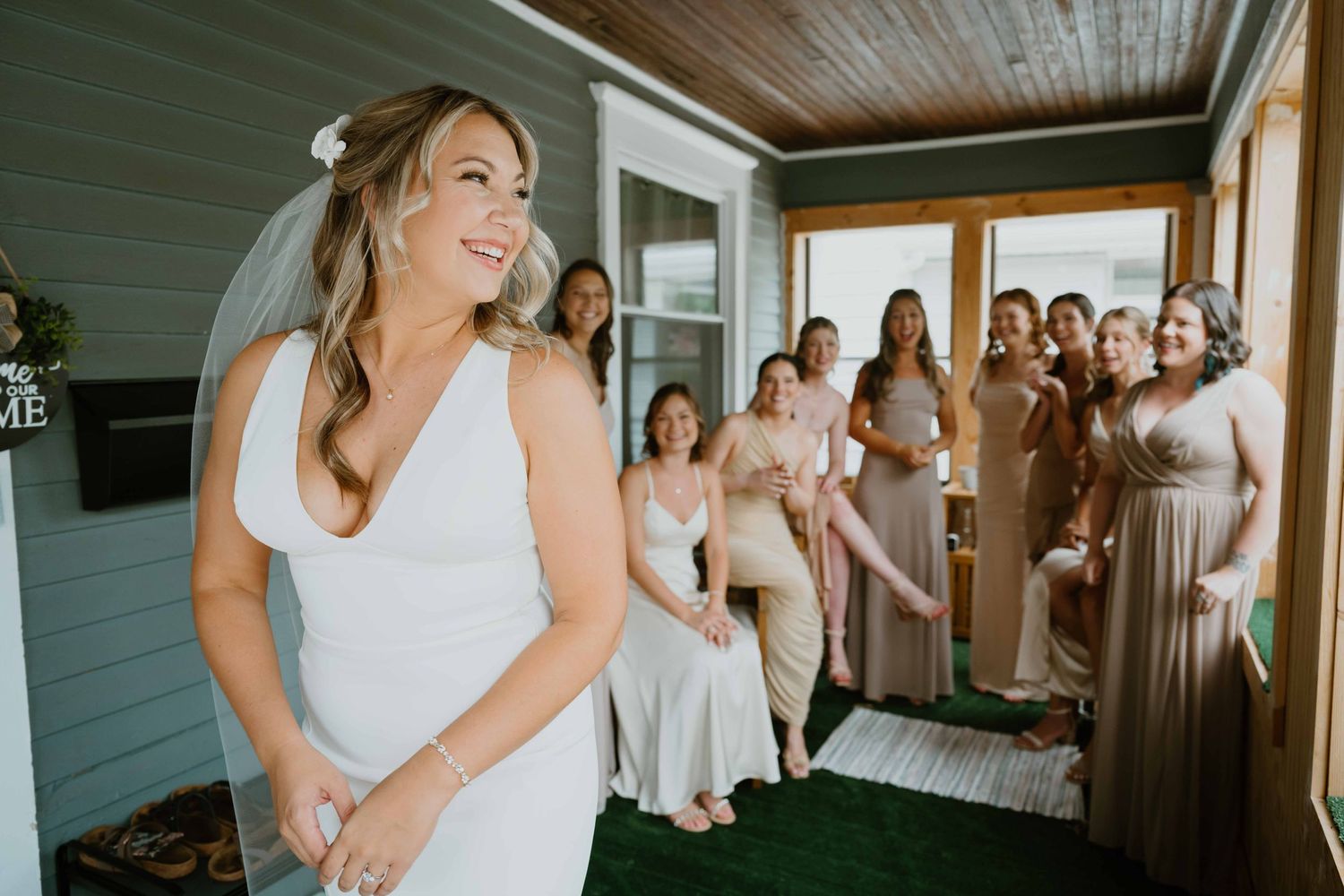 Bride in fitted white gown laughs joyfully during first look with cheering bridesmaids behind her.