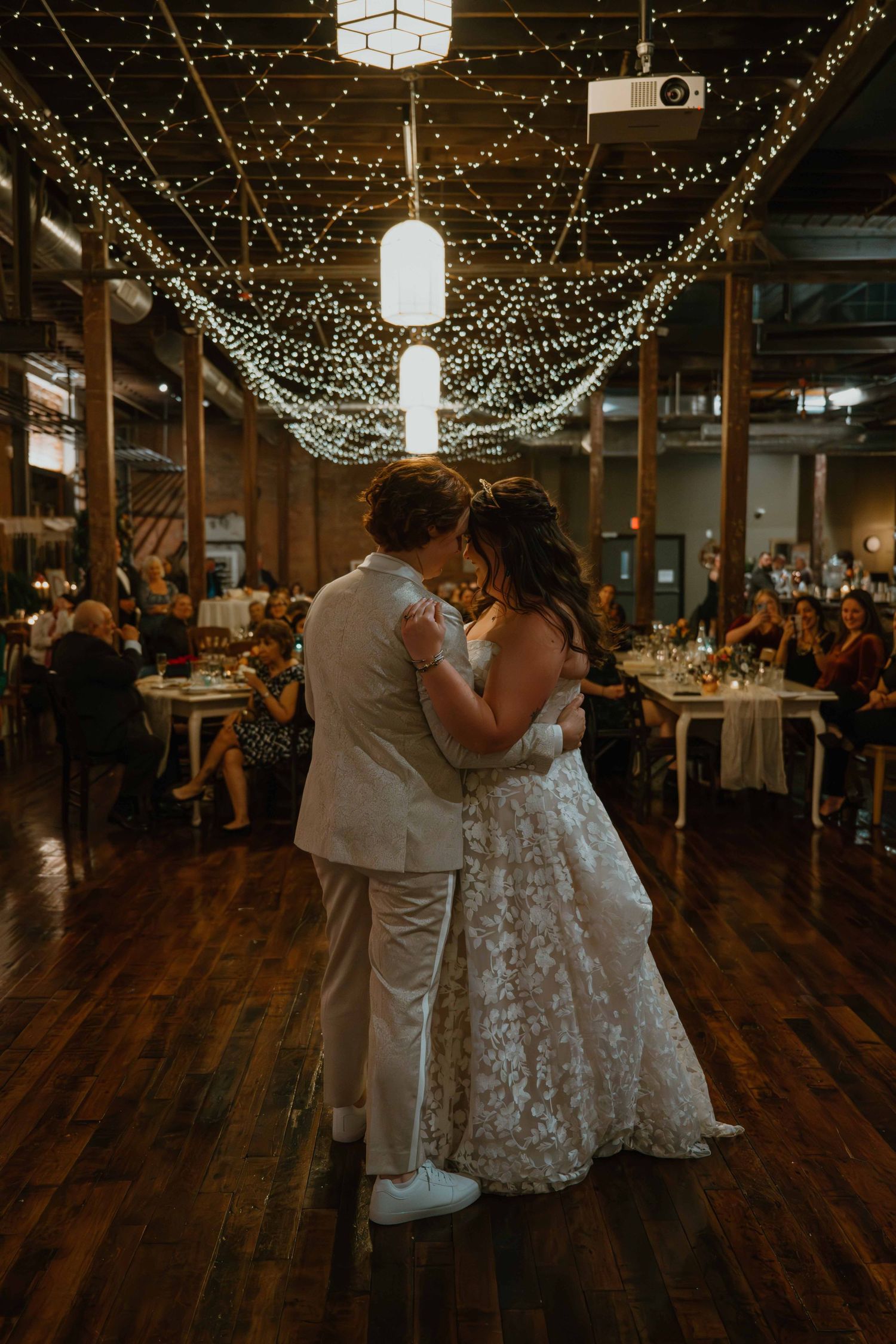 Couple shares first dance under twinkling fairy lights at elegant industrial wedding venue.