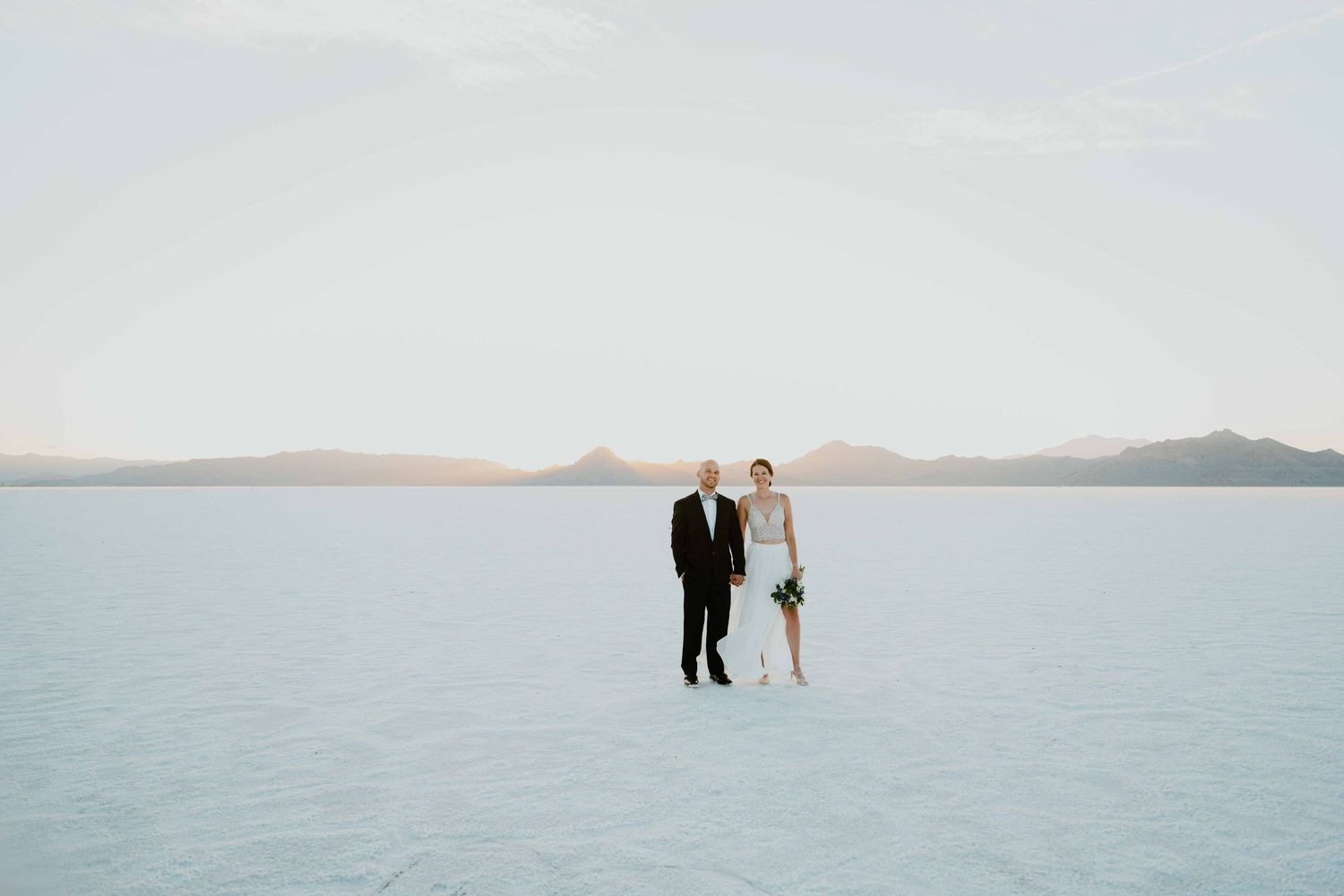 Bride and groom stand together on the vast white Bonneville Salt Flats with mountains in the background.