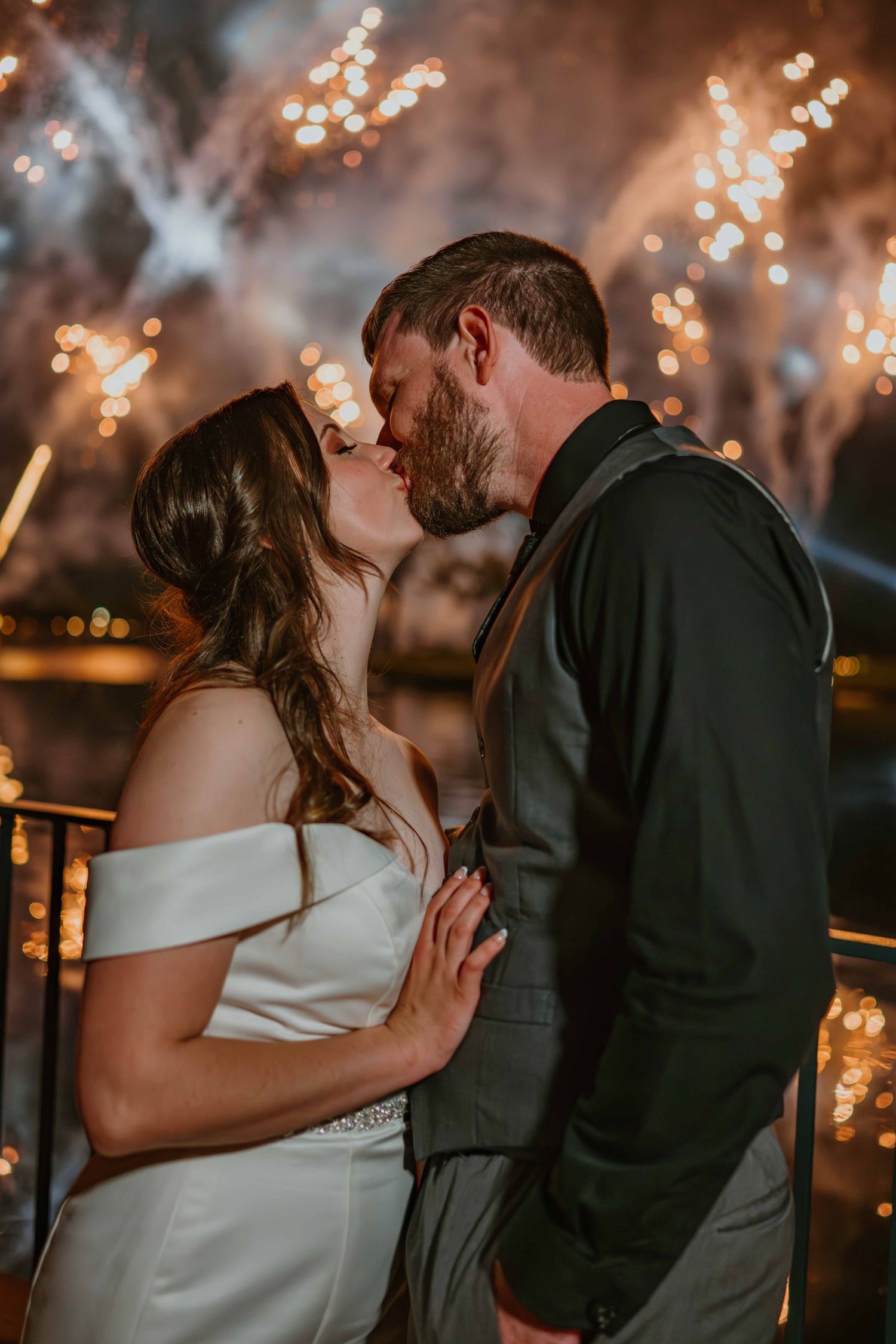 Bride and groom sharing romantic kiss with golden fireworks exploding behind them at night.