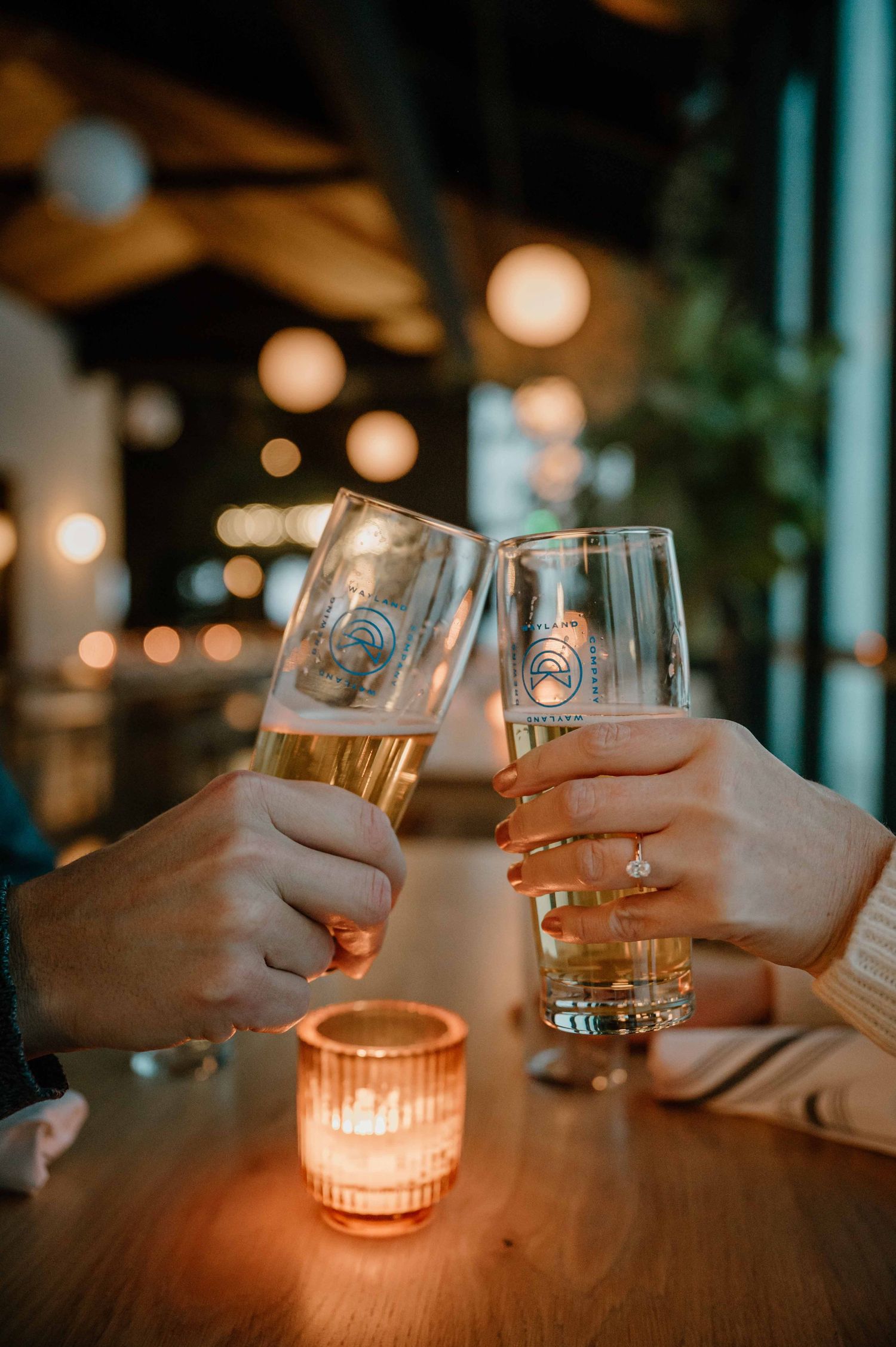 Two people clinking beer glasses over candlelit table in warmly lit restaurant during evening.