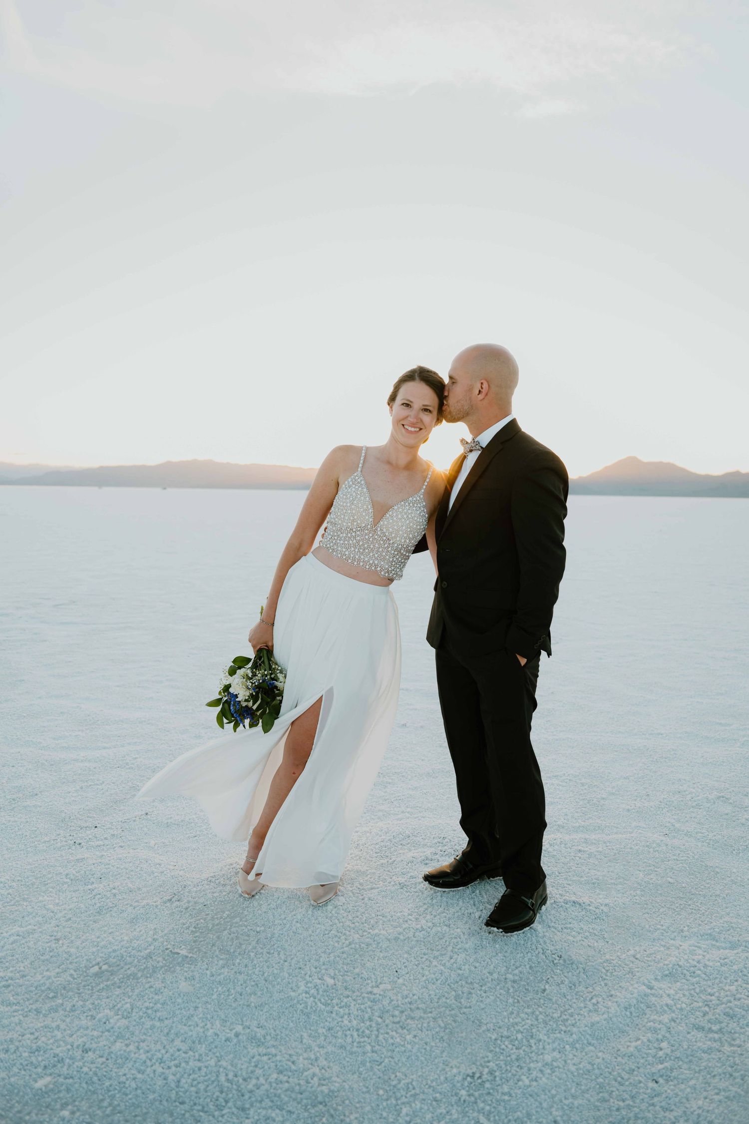 Groom kisses bride on cheek as she smiles holding a bouquet on the salt flats at sunset.