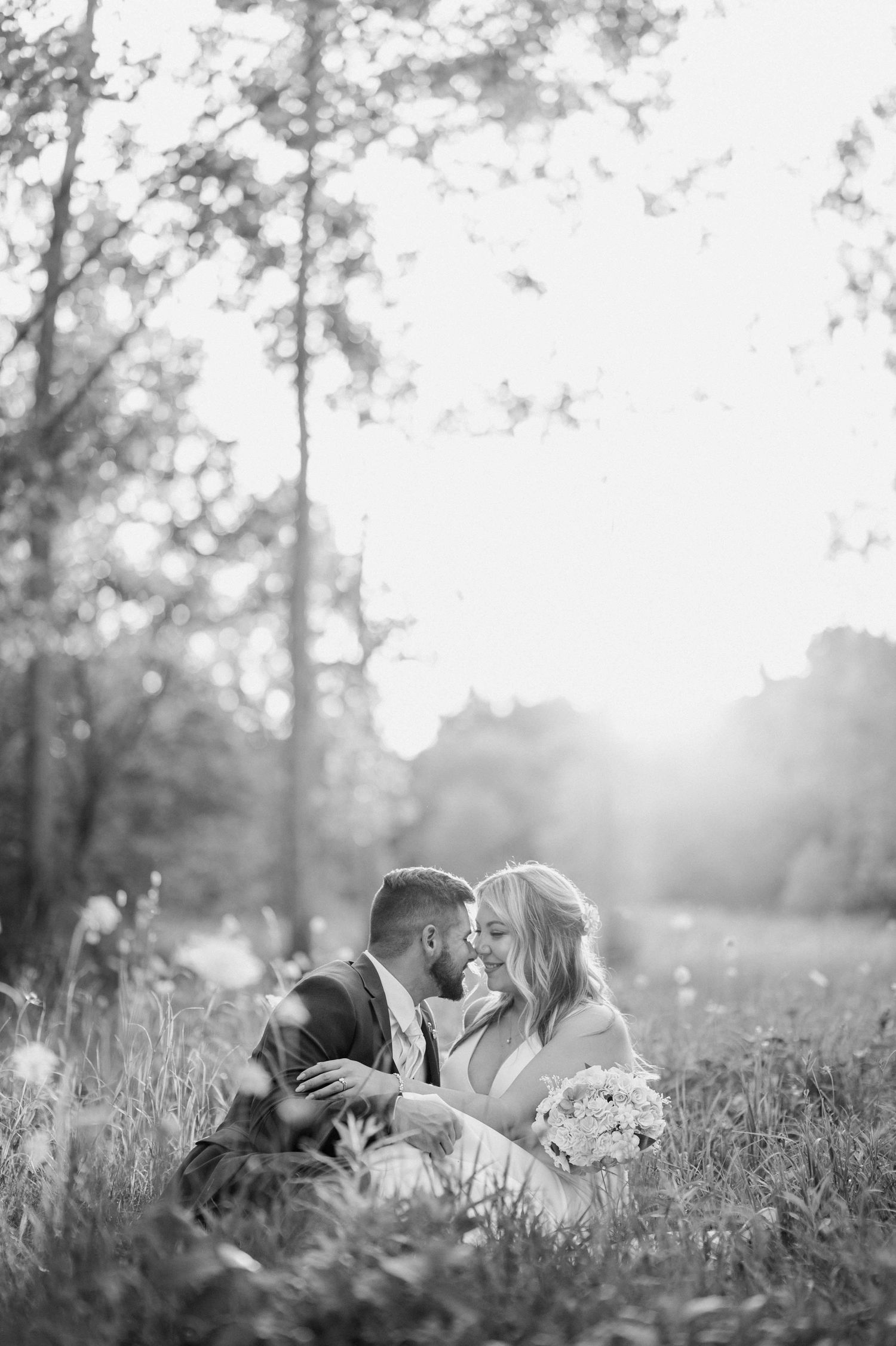 Romantic black and white photo of bride and groom sitting together in a sunlit garden meadow.