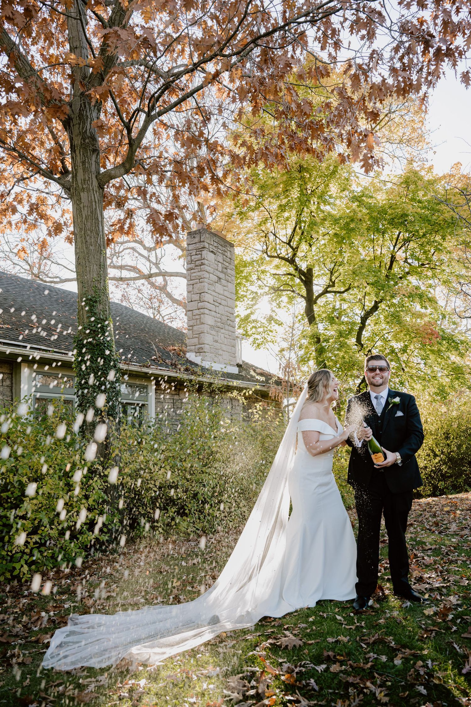 Bride and groom pop champagne bottle in autumn garden with colorful fall foliage backdrop.