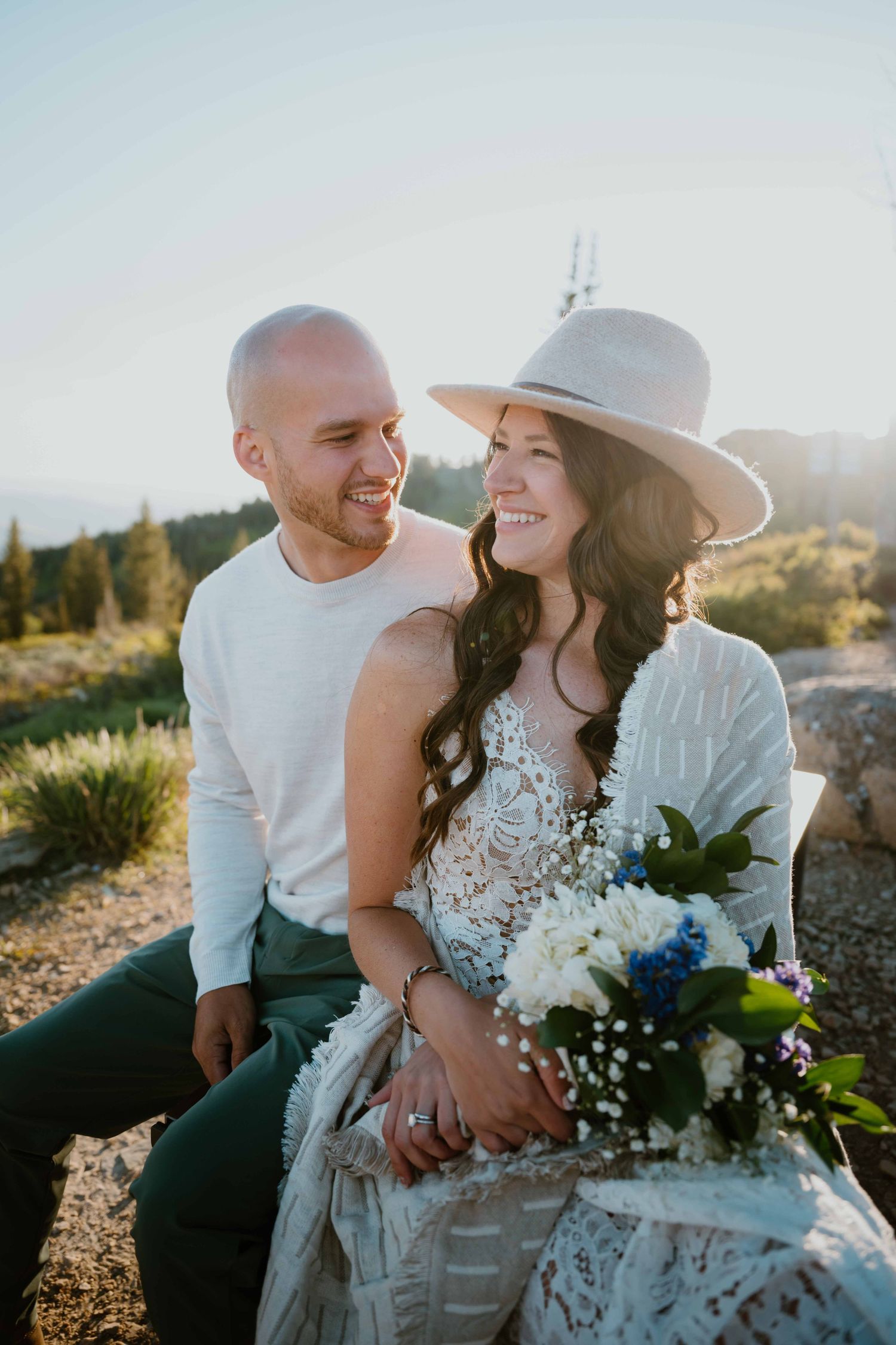 Smiling couple sits together at golden hour, bride in boho lace dress and wide-brim hat with bouquet.