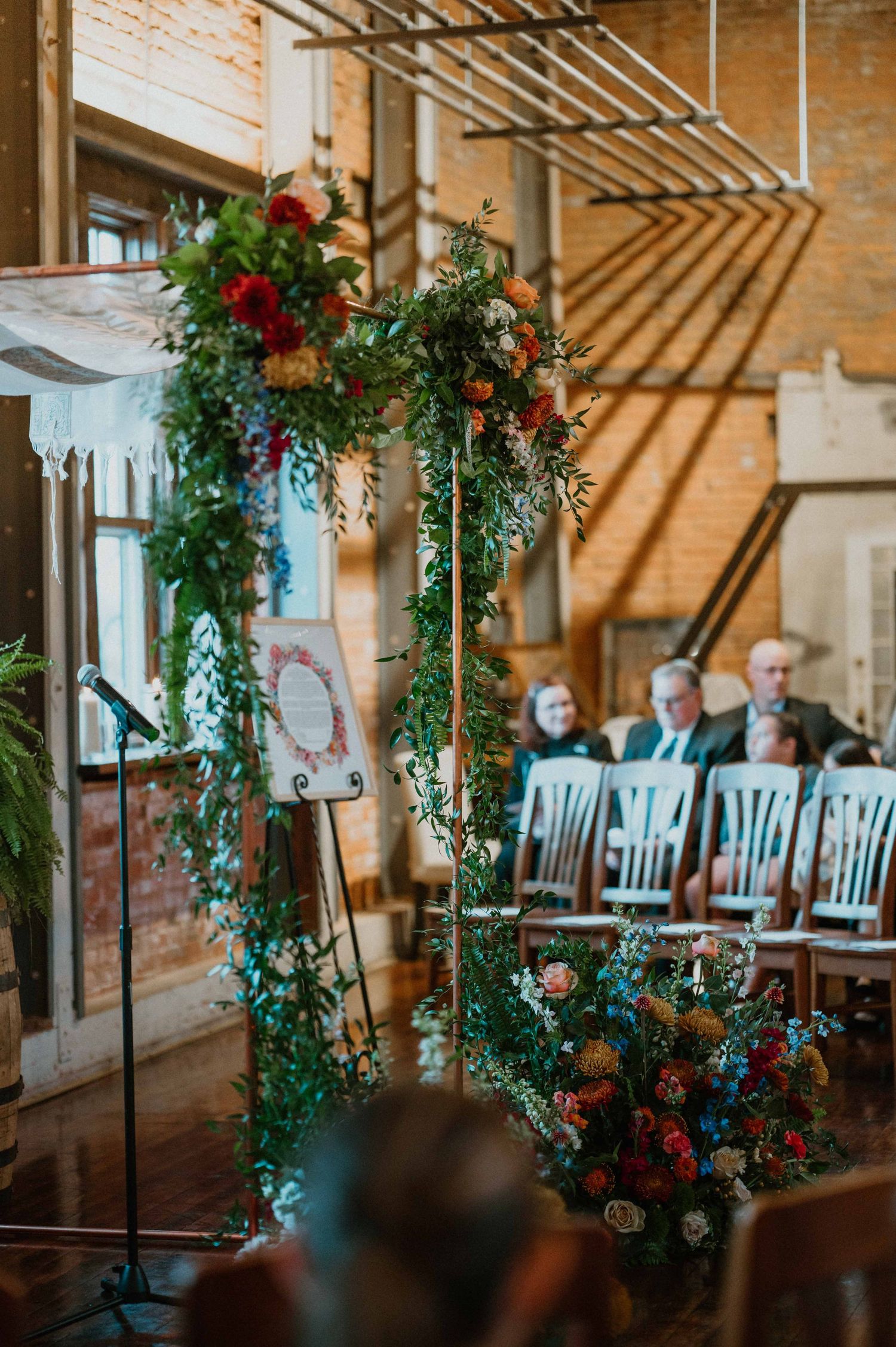 Floral wedding arch with greenery and colorful blooms inside rustic brick ceremony venue.