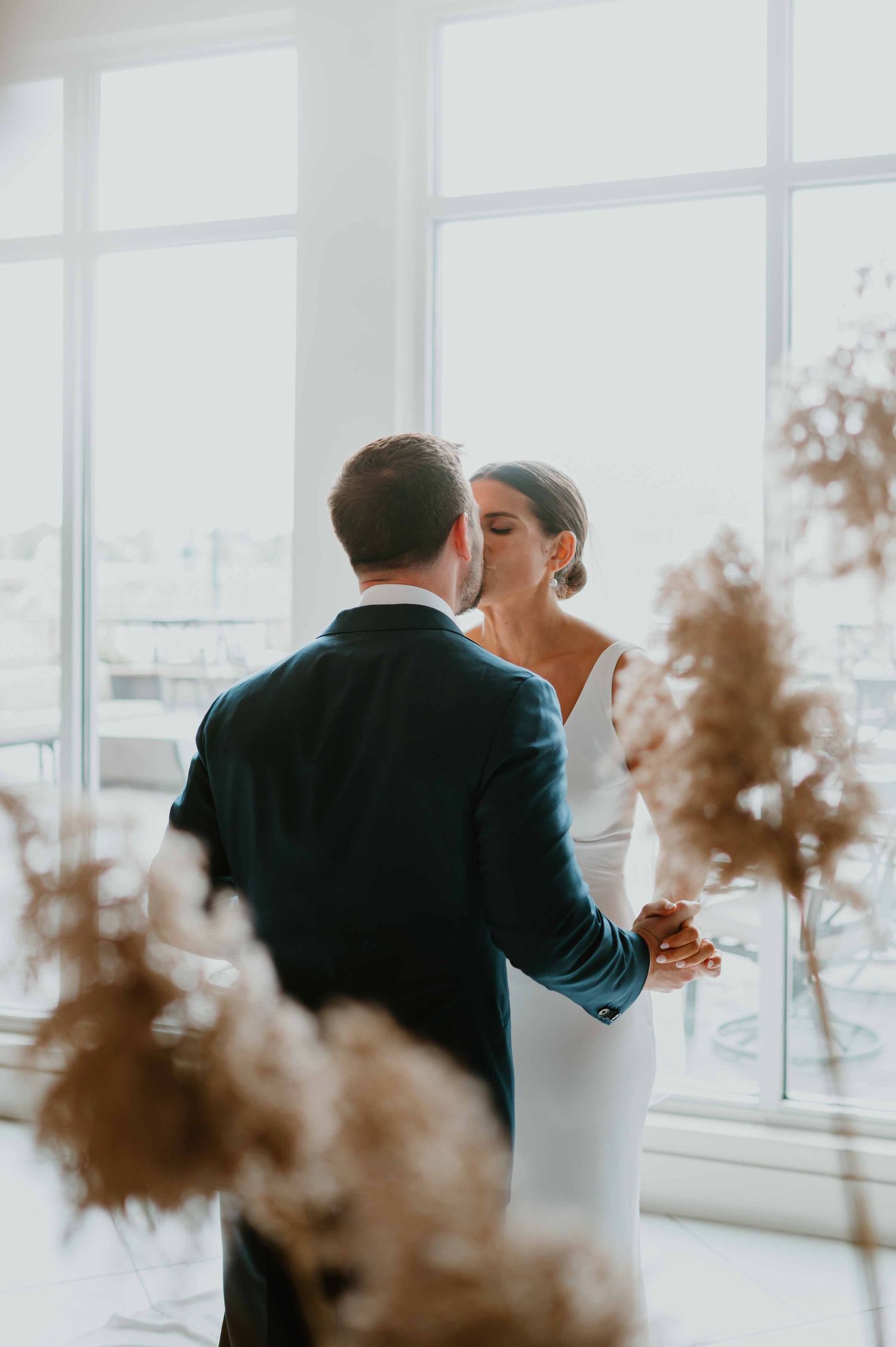 Bride and groom dance near bright windows surrounded by pampas grass.
