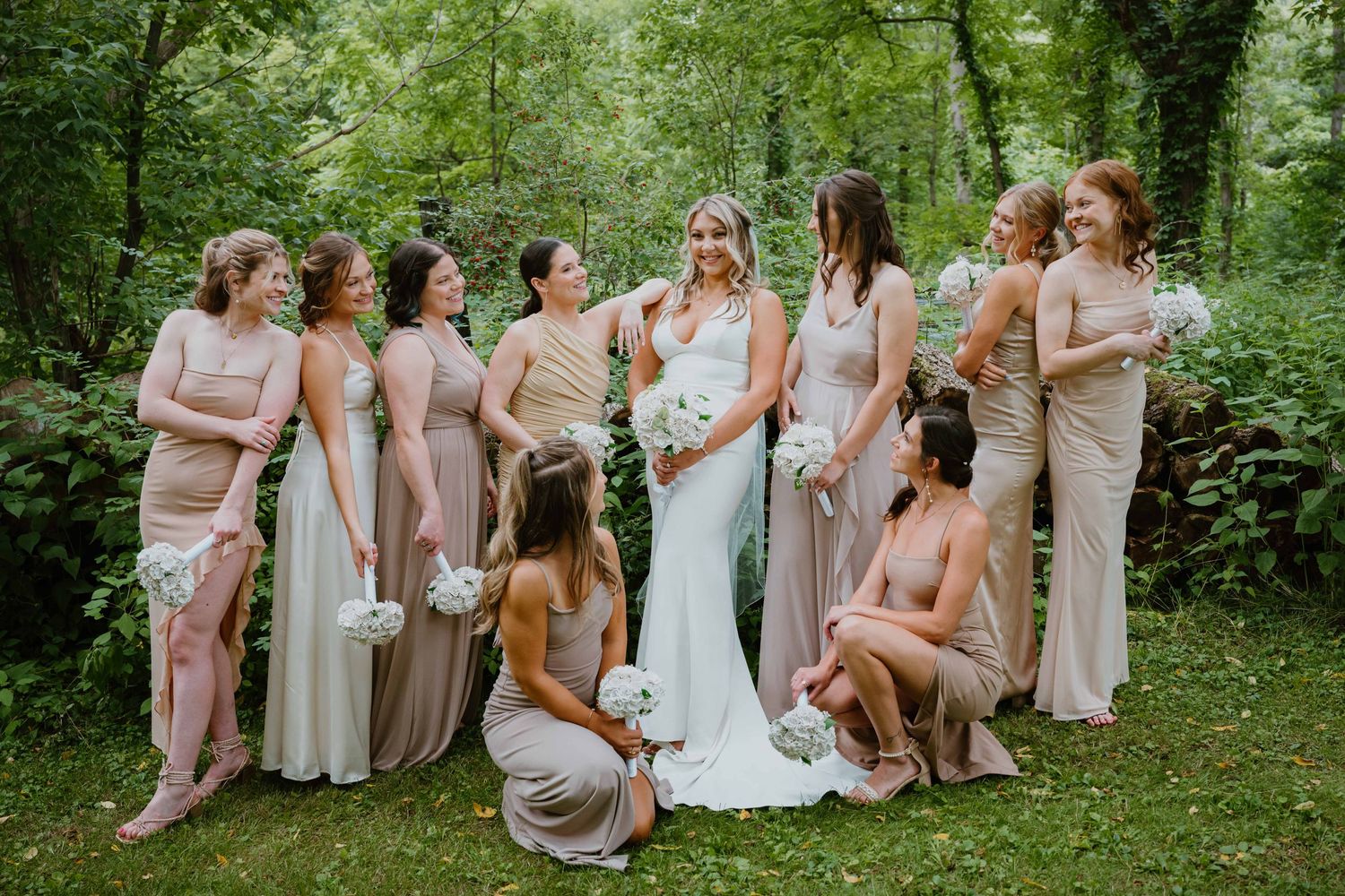 Bride with bouquet poses with bridesmaids in blush and champagne gowns in lush garden setting.
