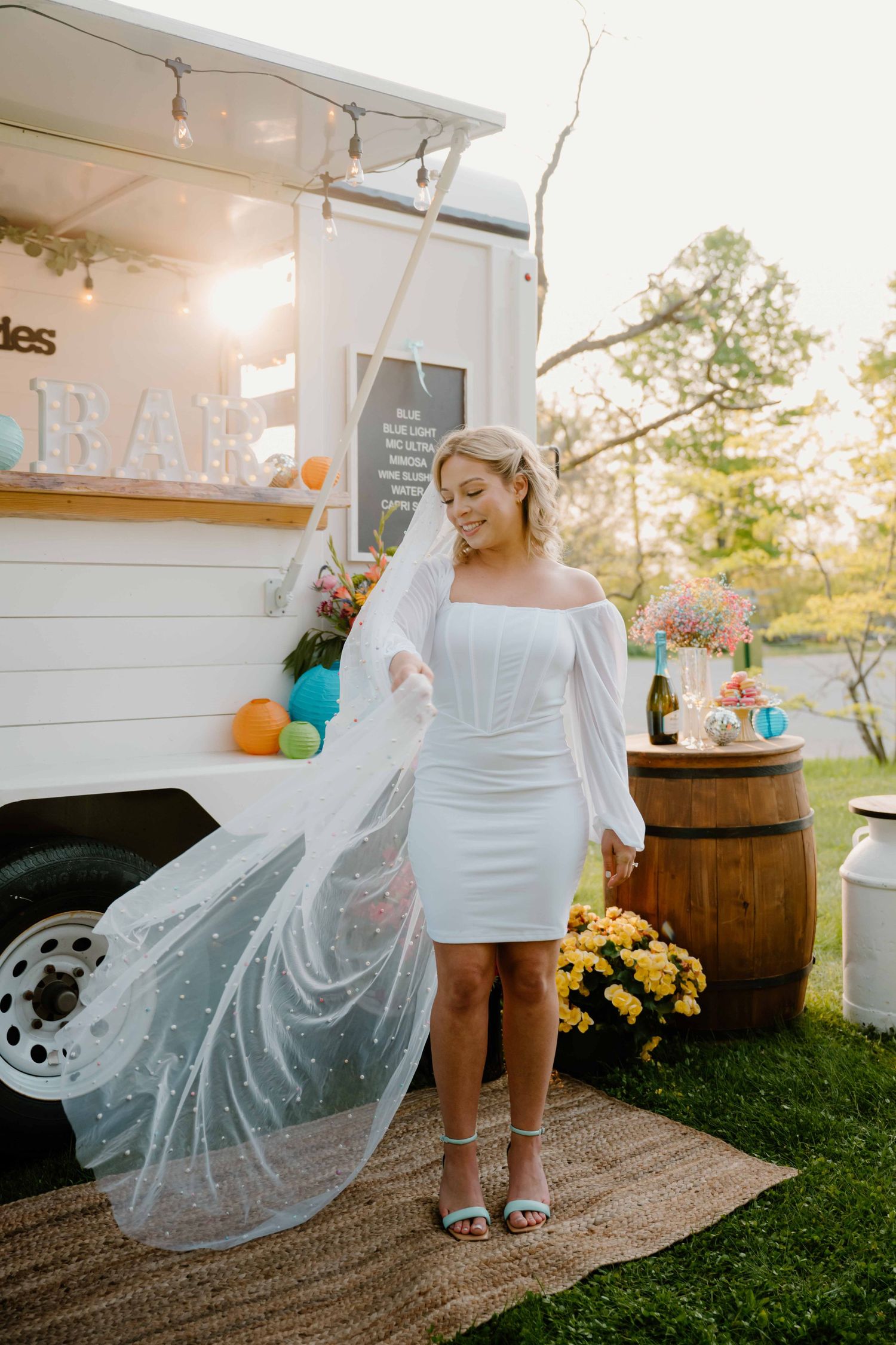 Bride in short white dress and polka dot veil poses by a white bar truck with colorful balloons outdoors.