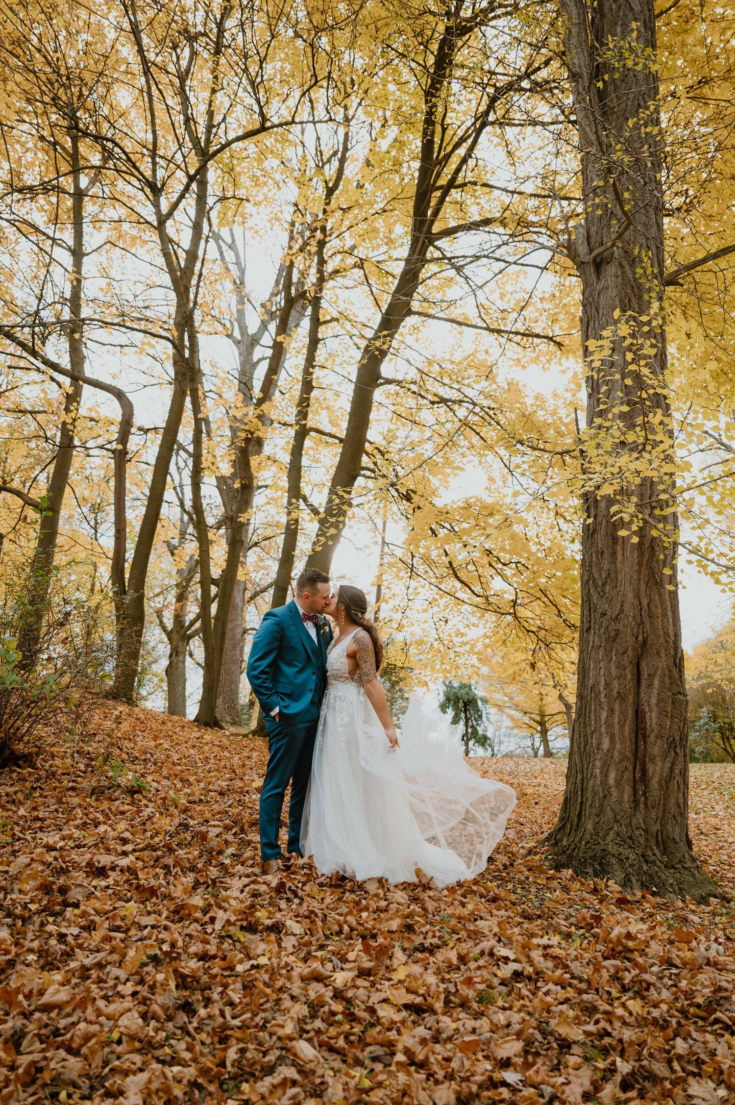 Bride and groom kiss surrounded by golden fall foliage and leaf-covered ground in autumn forest.