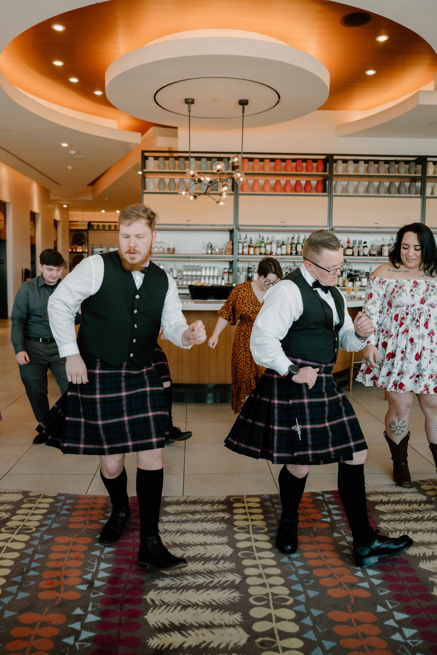 Men in Scottish kilts performing traditional Highland dance at wedding reception near hotel bar.