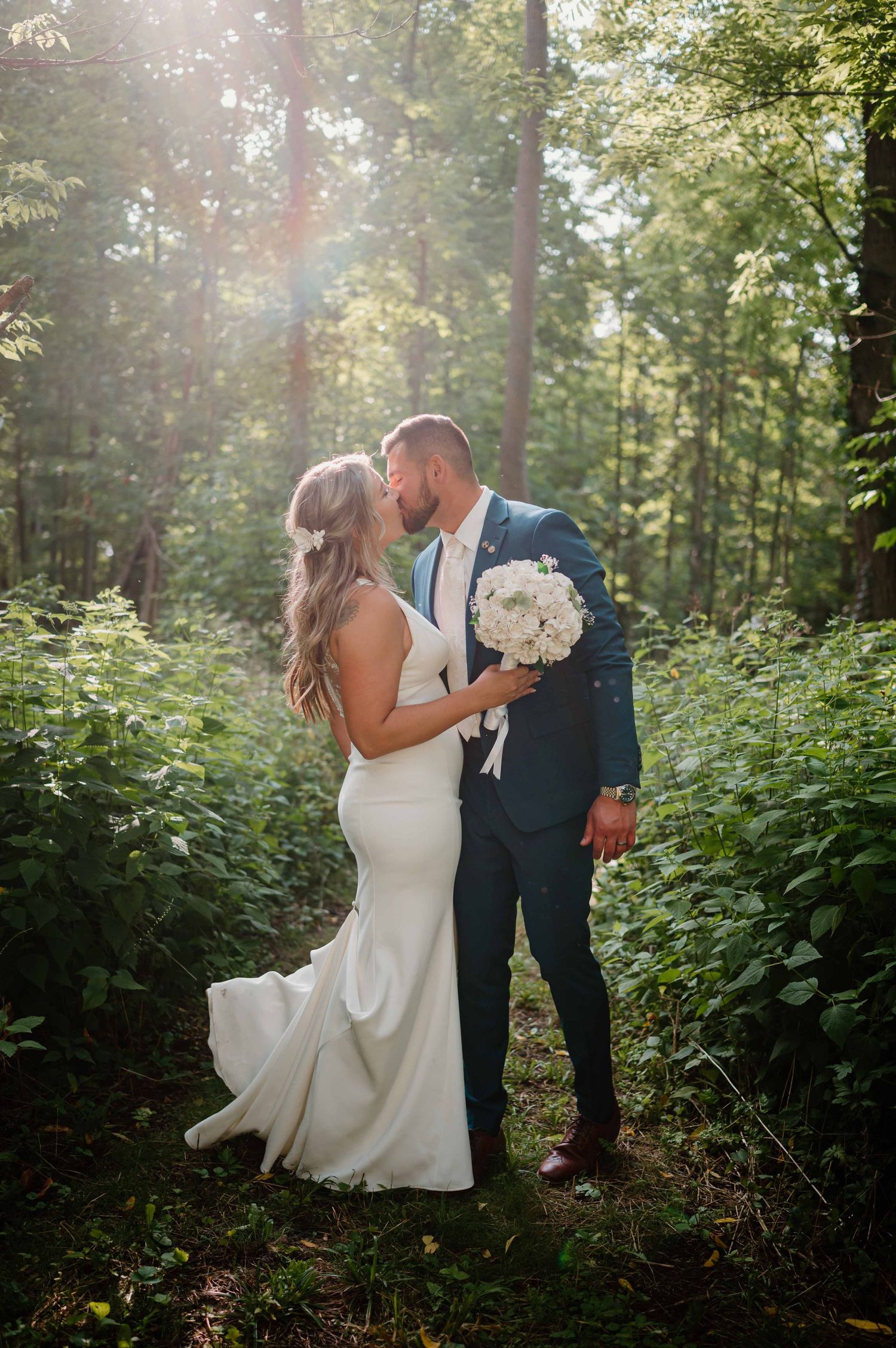 Bride and groom kissing in a lush green forest with warm sunlight streaming through the trees.