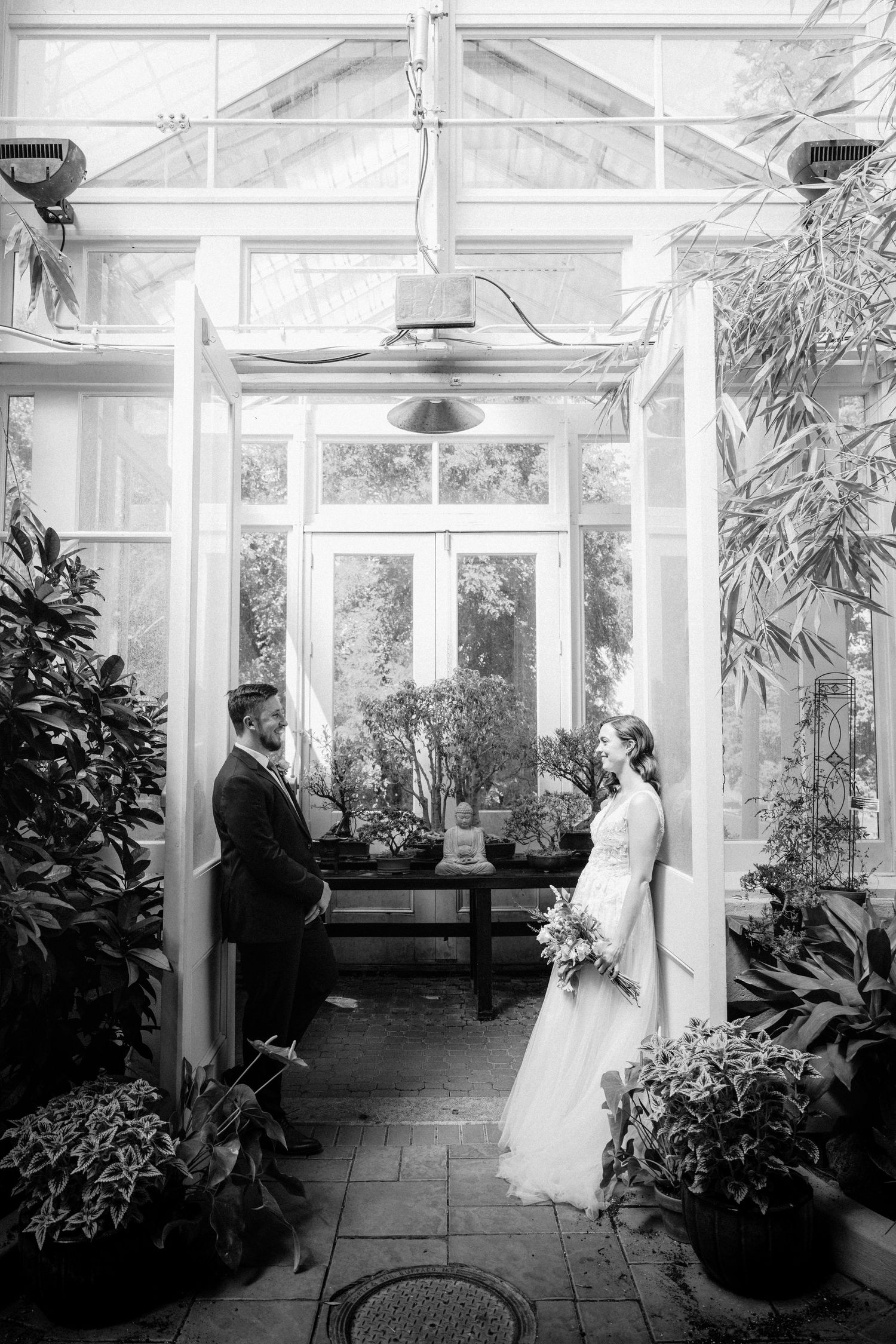 Black and white photo of bride and groom facing each other inside a sunlit glass greenhouse.