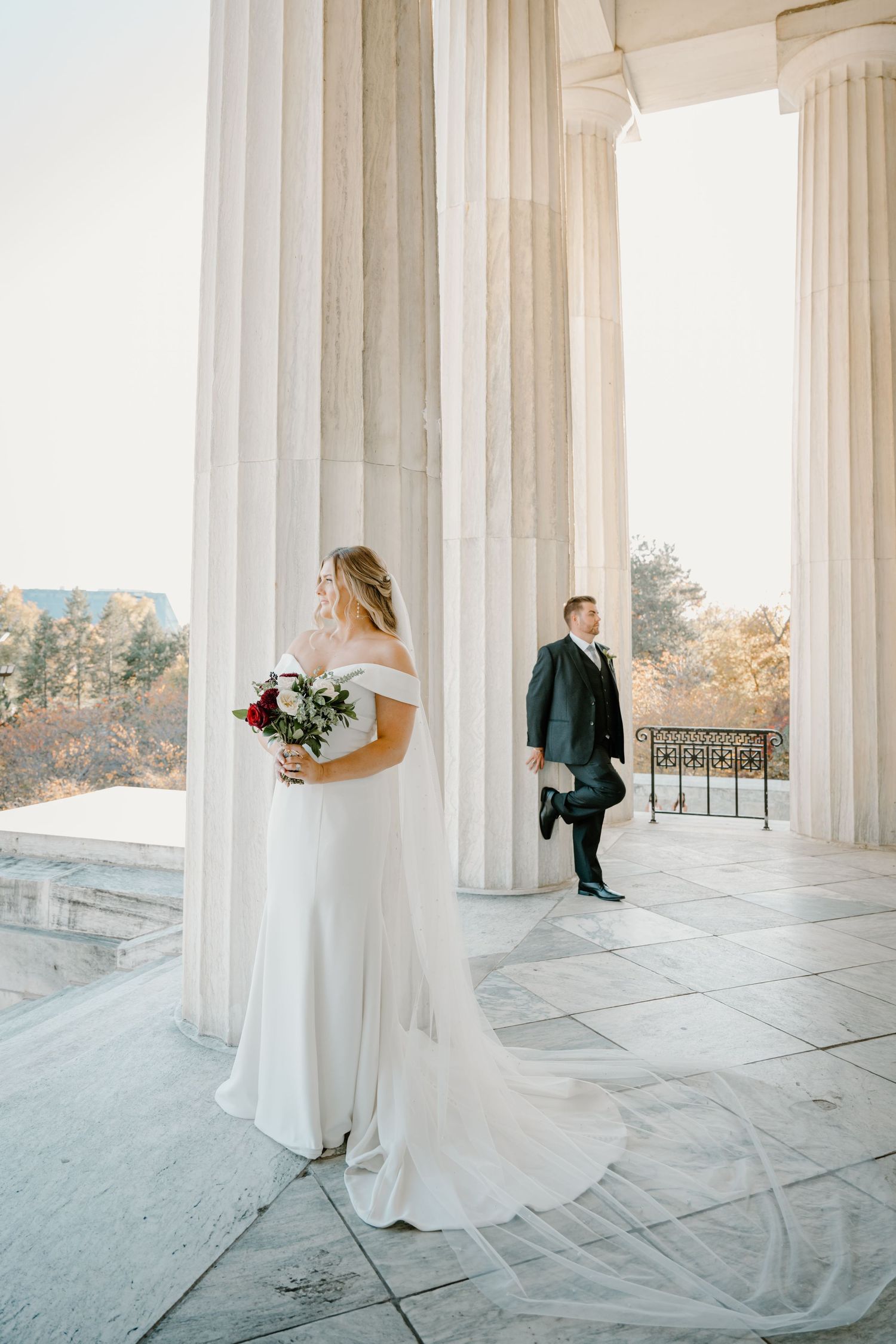 Bride in off-shoulder gown holding red bouquet stands before grand white columns as groom waits in background.